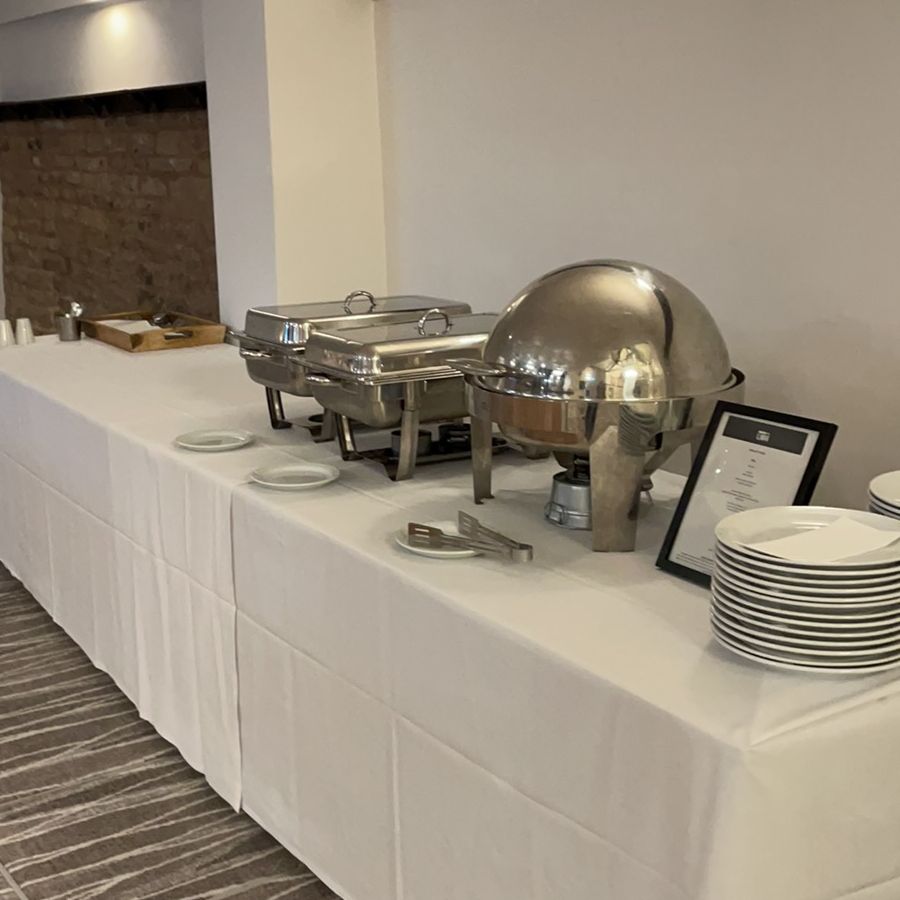 Buffet station set up in a conference room with serving trays, plates, and utensils on a long table covered with a white cloth.
