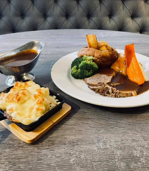 A plated Sunday roast meal with sliced roast beef, potatoes, broccoli, carrot, gravy, a serving of cheesy cauliflower bake, and a gravy boat.