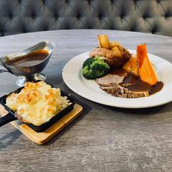 A plated Sunday roast meal with sliced roast beef, potatoes, broccoli, carrot, gravy, a serving of cheesy cauliflower bake, and a gravy boat.
