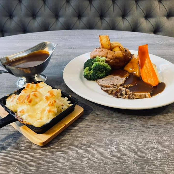 A plated Sunday roast meal with sliced roast beef, potatoes, broccoli, carrot, gravy, a serving of cheesy cauliflower bake, and a gravy boat.