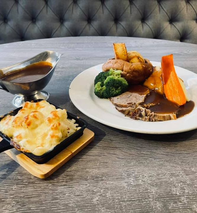 A plated Sunday roast meal with sliced roast beef, potatoes, broccoli, carrot, gravy, a serving of cheesy cauliflower bake, and a gravy boat.