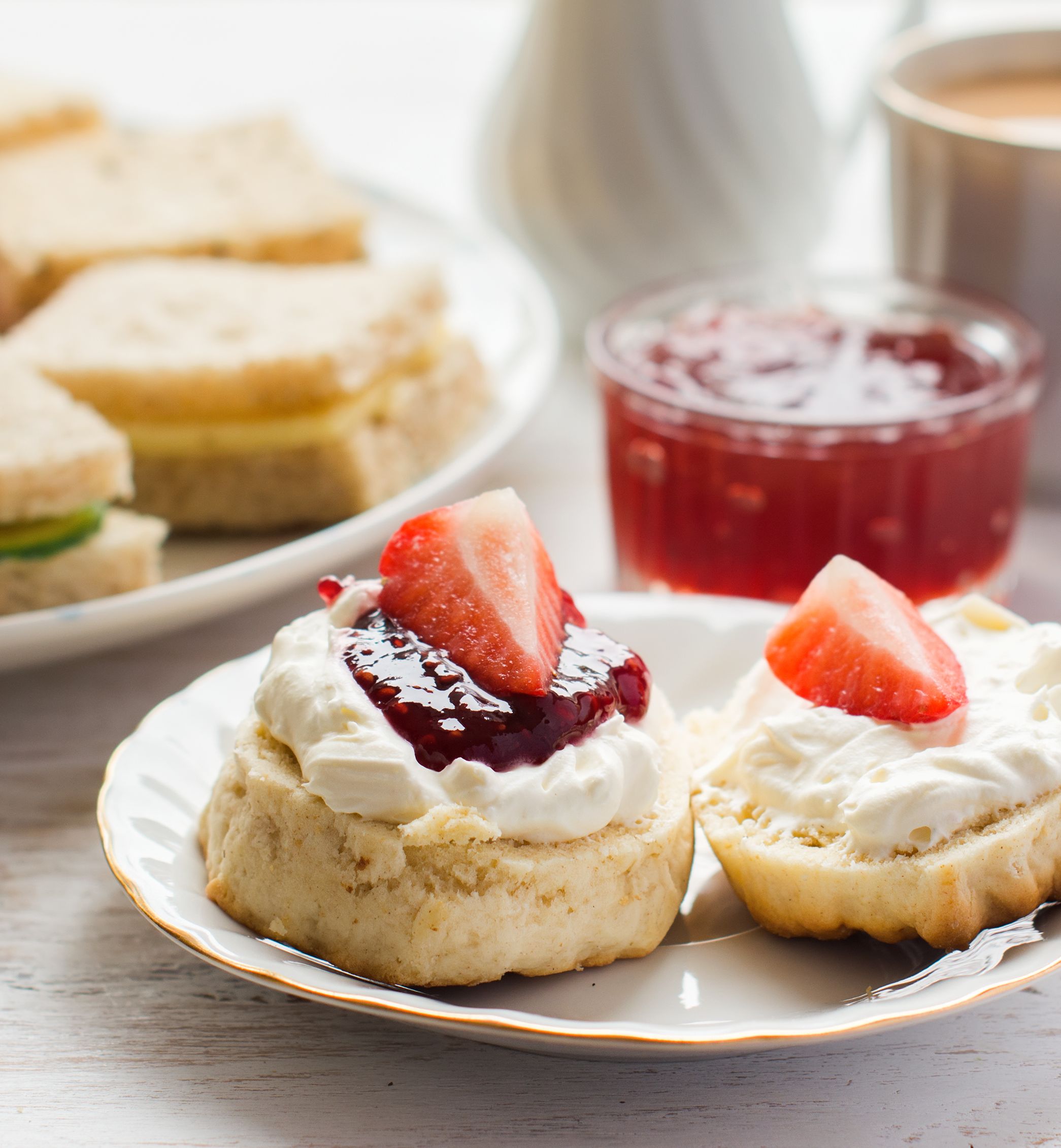 Afternoon tea with scones topped with cream, jam, and strawberries, alongside assorted finger sandwiches and a cup of tea.