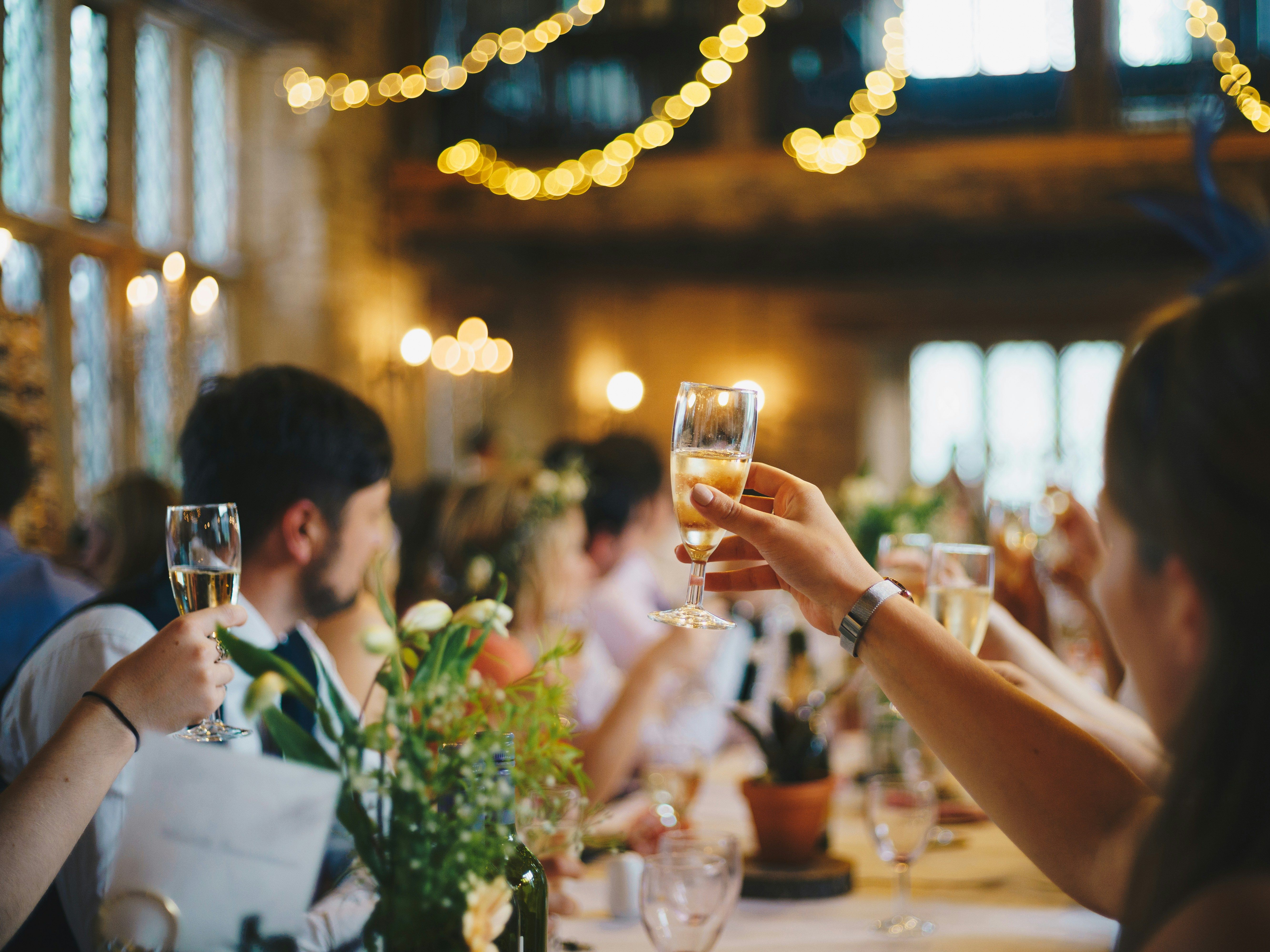 People raising champagne glasses in a celebratory toast at a festive indoor event with string lights in the background.