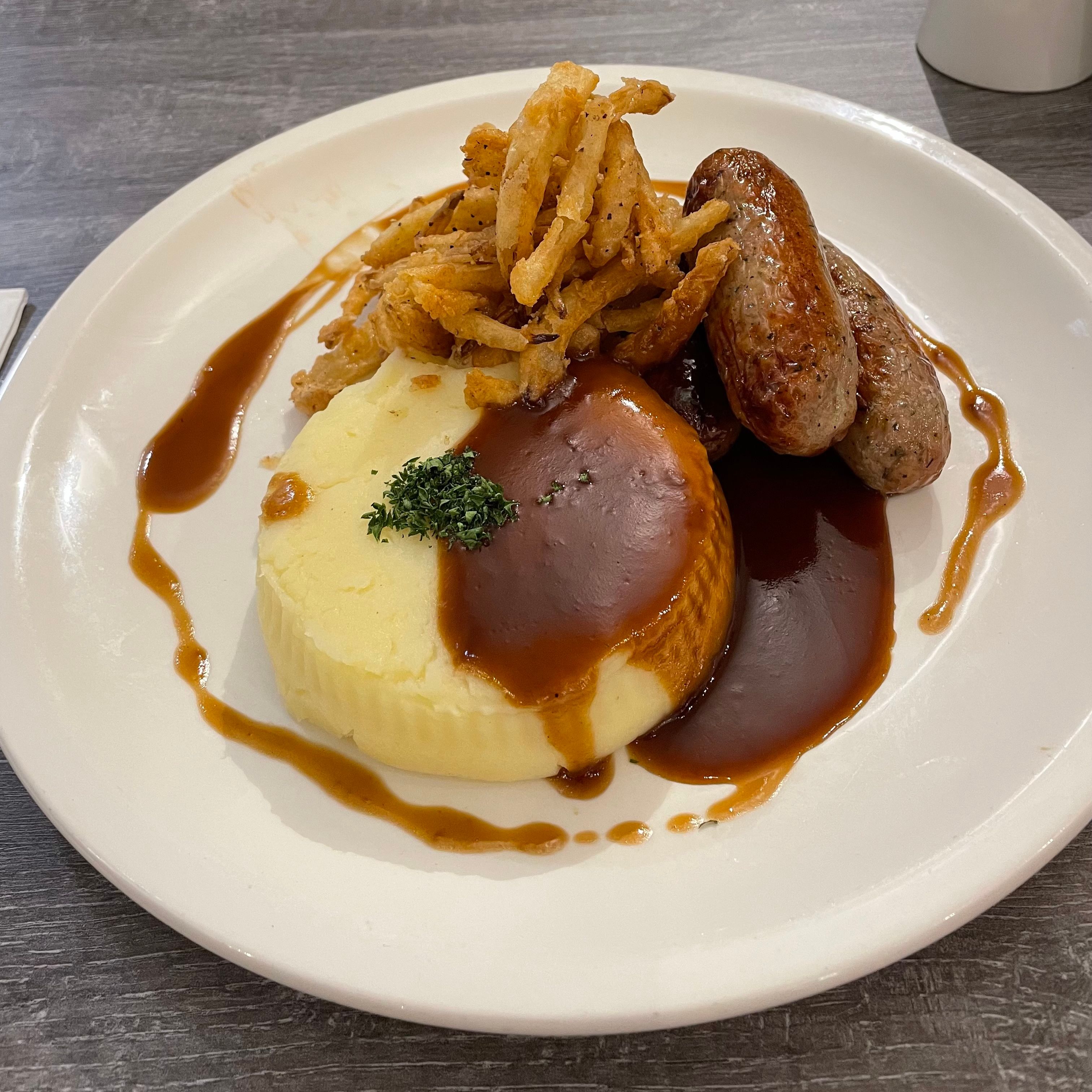 A plate with mashed potatoes, sausages, crispy fried onions, and brown gravy.