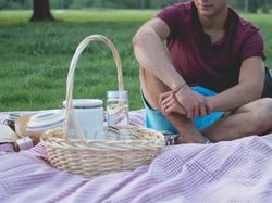 Person sitting on a picnic blanket with a picnic basket and jars on grass