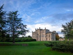 Historic stone mansion with large windows surrounded by lush green garden and tall trees under a blue sky.
