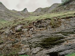 A child in a yellow jacket stands near the entrance of a rocky cave at the base of a grassy hill.