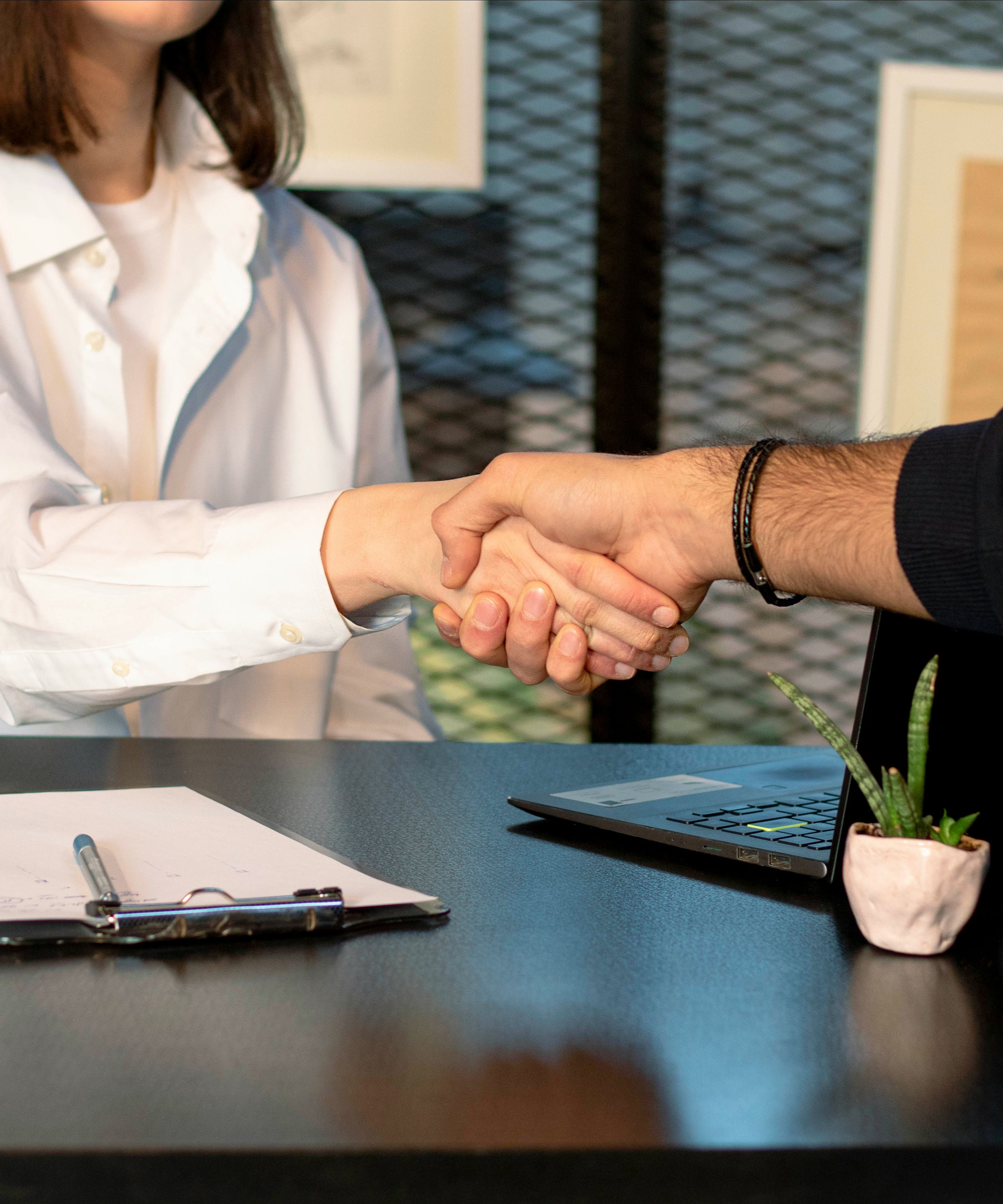 Two people shaking hands across a desk with a clipboard, laptop, and small plant