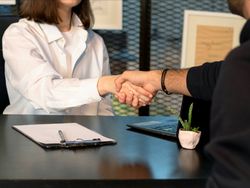 Two people shaking hands across a desk with a clipboard, laptop, and small plant