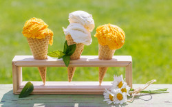 A trio of ice cream cones—yellow and white—neatly arranged in a wooden stand with fresh mint leaves and a bunch of daisies on a green outdoor surface.