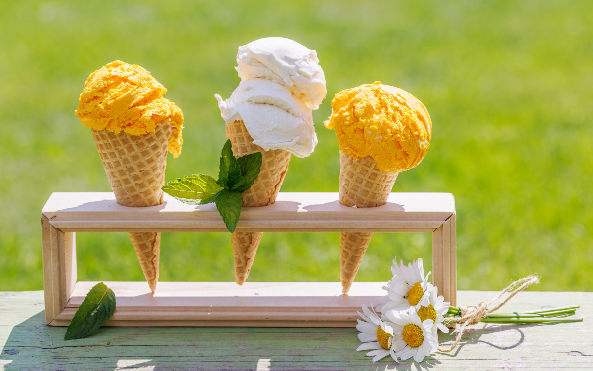 A trio of ice cream cones—yellow and white—neatly arranged in a wooden stand with fresh mint leaves and a bunch of daisies on a green outdoor surface.