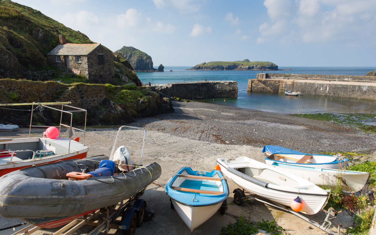 A peaceful harbor with several small boats on land, a stone building on a grassy hill, and calm water bordered by rock formations.