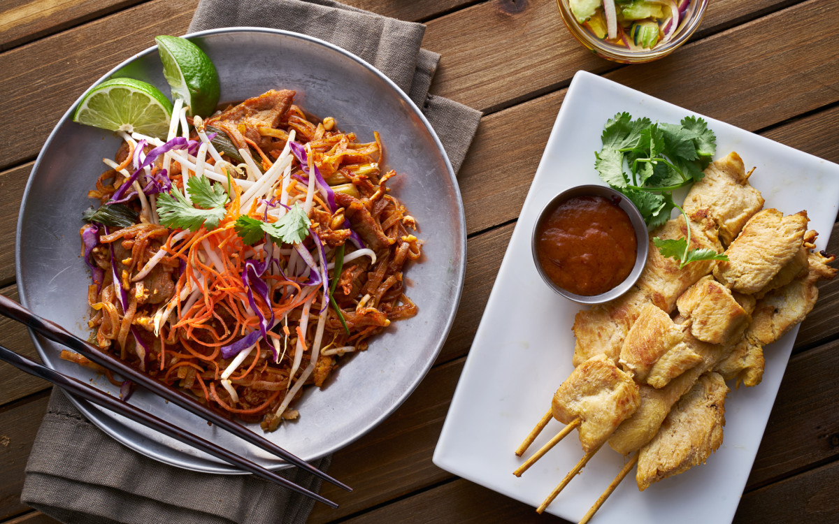 A close-up of pad thai noodles with vibrant garnishes next to a plate of chicken skewers served with a bowl of dipping sauce, placed on a wooden dining table.
