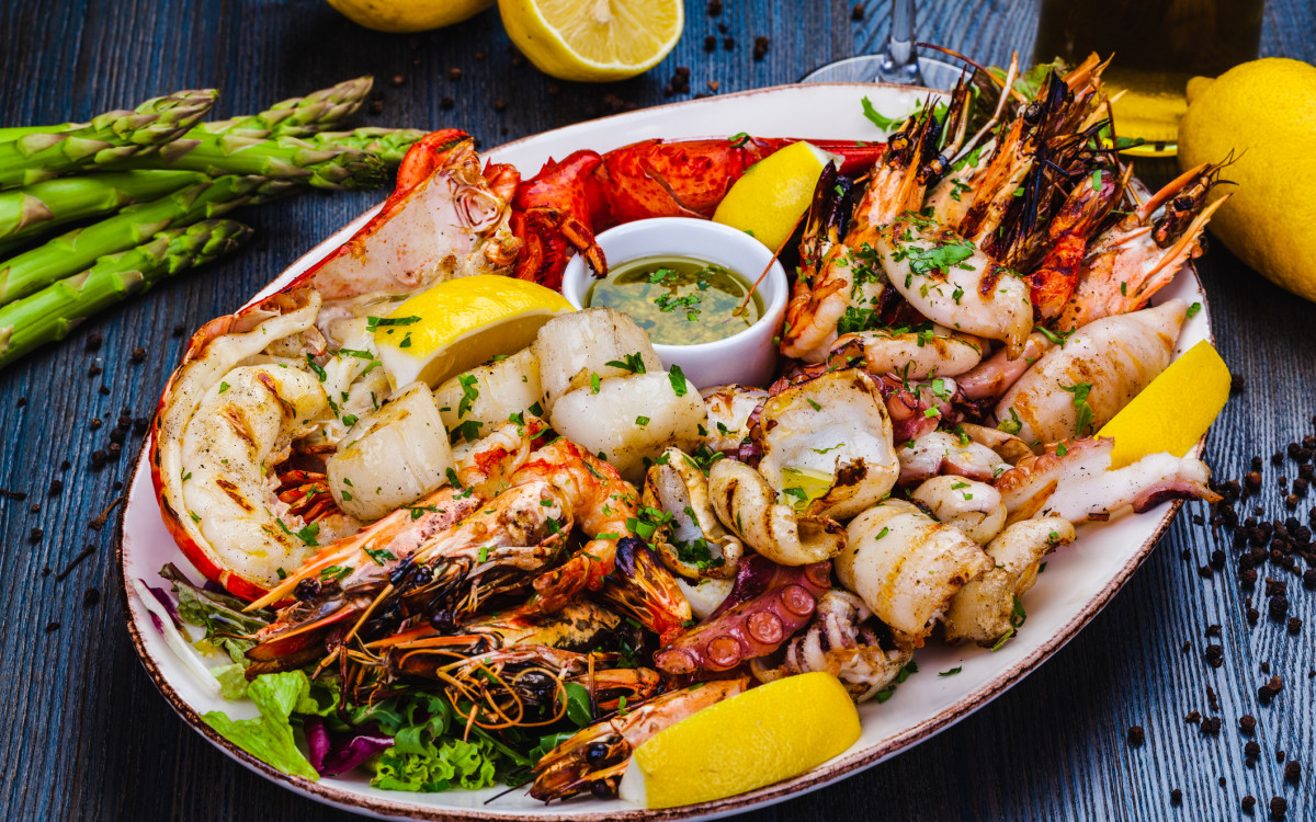 A plate of grilled seafood, including lobster, shrimp, and squid, accompanied by lemon wedges and a bowl of sauce, garnished with herbs on a wooden background.