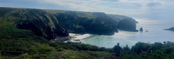 Coastal view with green cliffs, a rocky beach, and sunlight reflecting on the sea.
