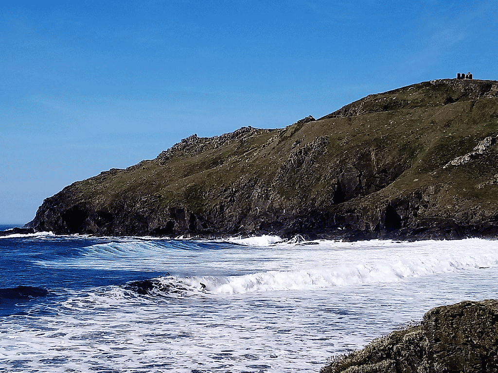 Rocky coastline with waves crashing against the shore and grassy hills under a blue sky.