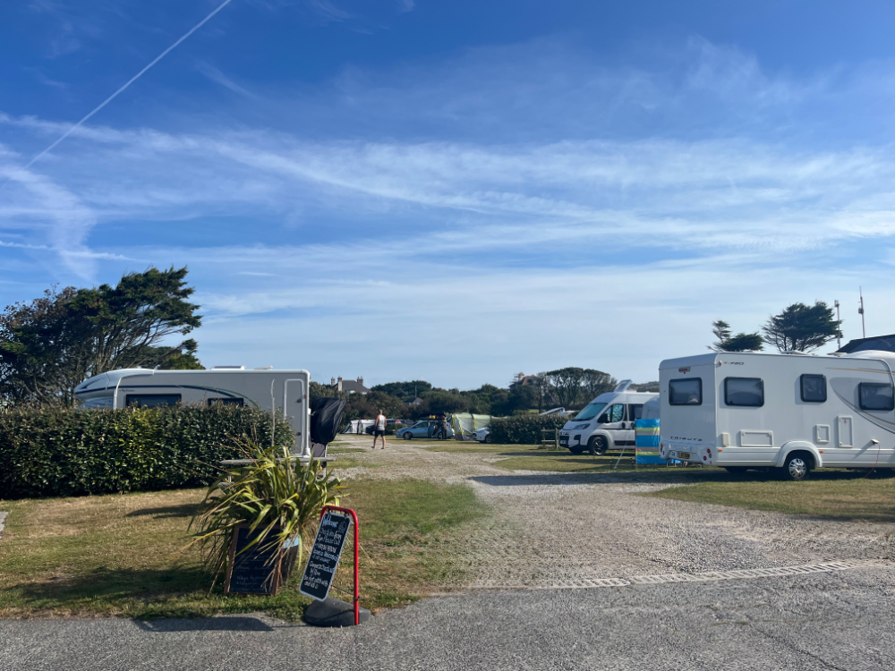 Campsite entrance with caravans and campervans parked on hardstanding pitches under a blue sky.