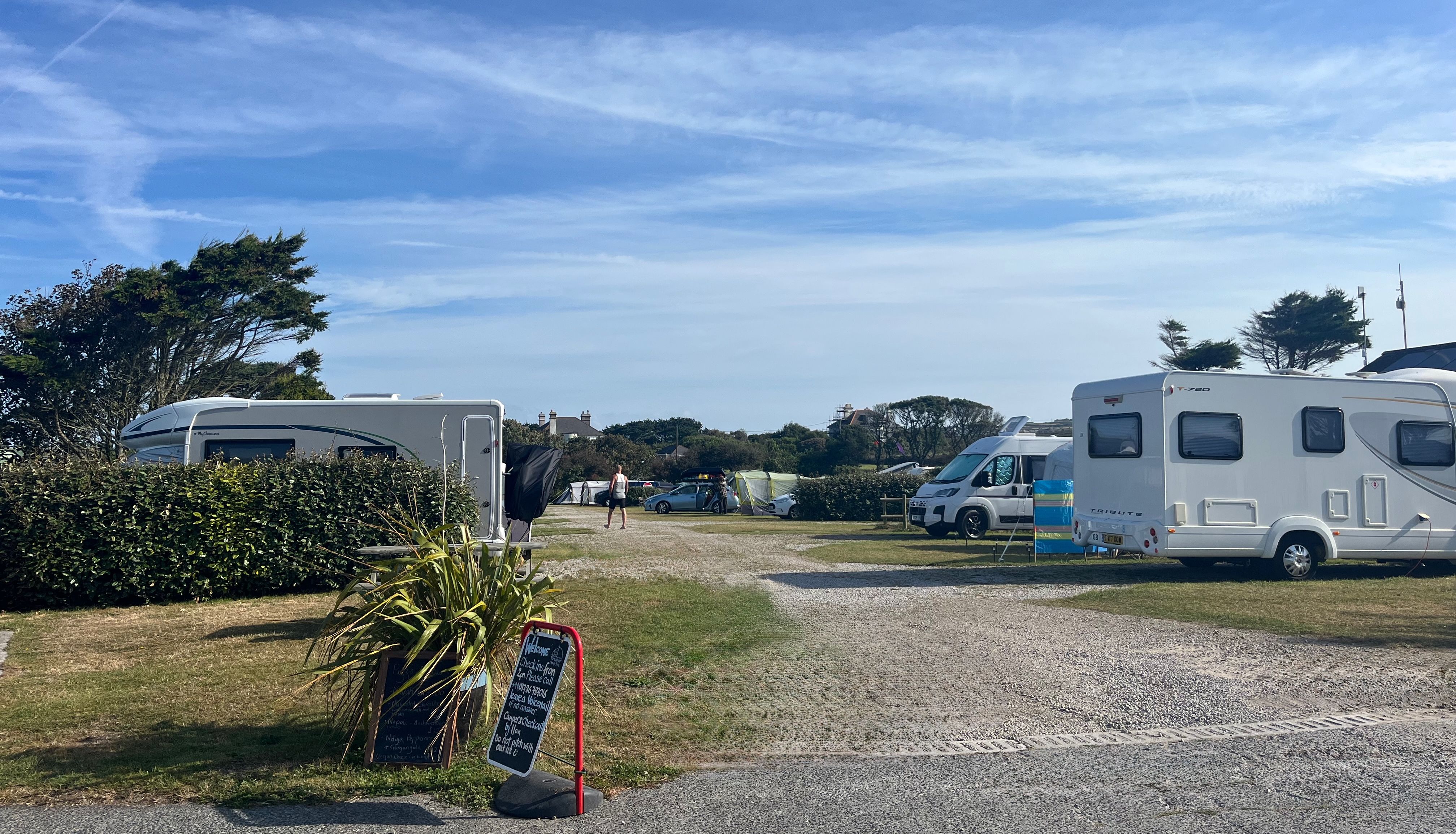 Campsite entrance with caravans and campervans parked on hardstanding pitches under a blue sky.