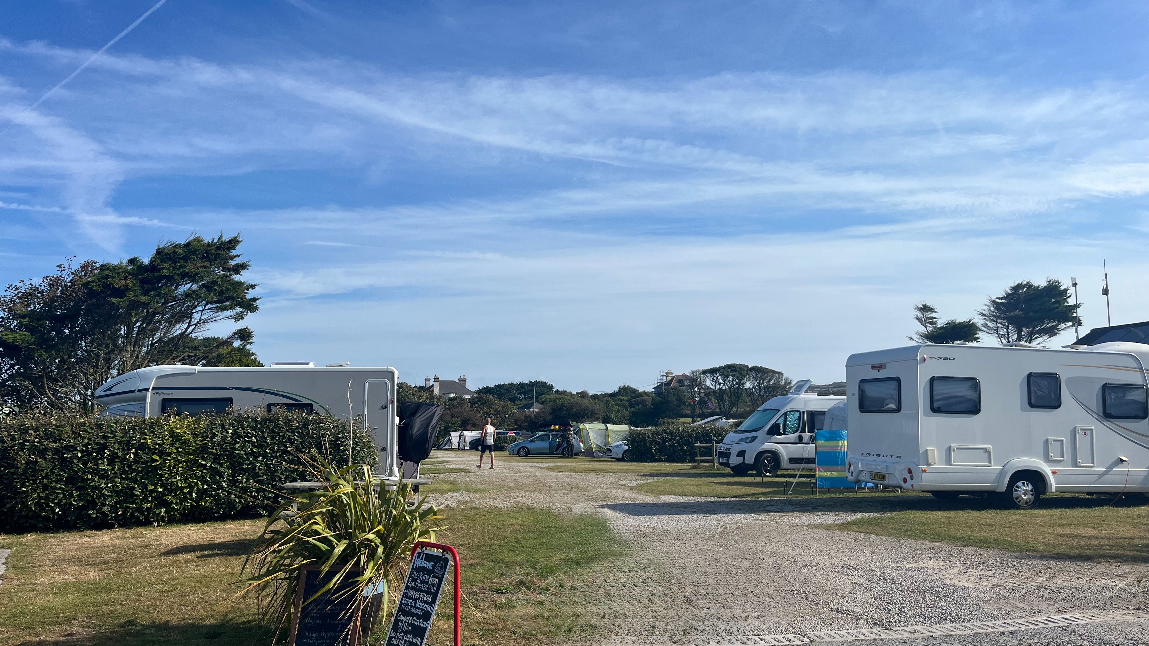 Campsite entrance with caravans and campervans parked on hardstanding pitches under a blue sky.