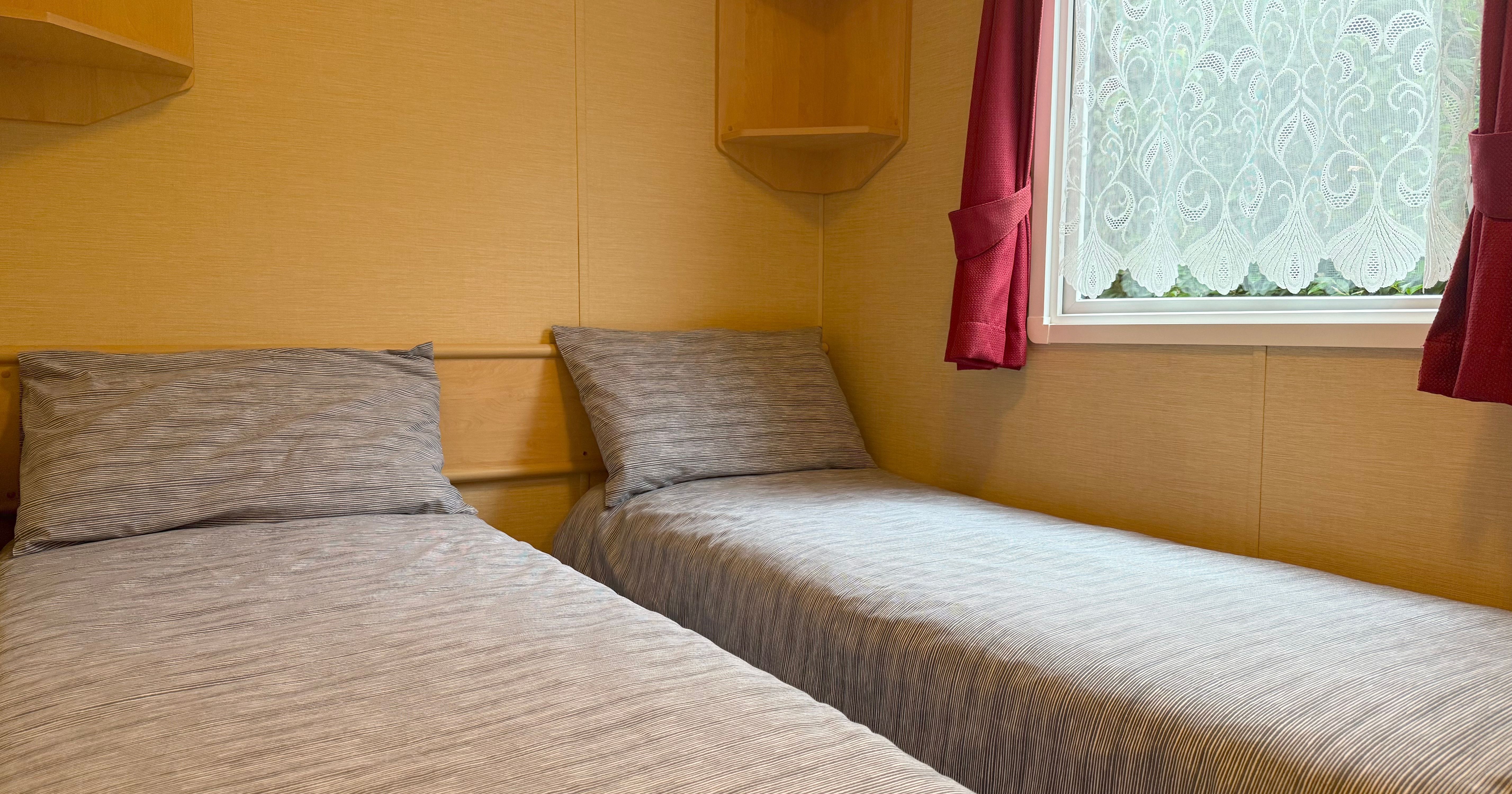 Two single beds with gray bedding in a small room with a window and red curtains