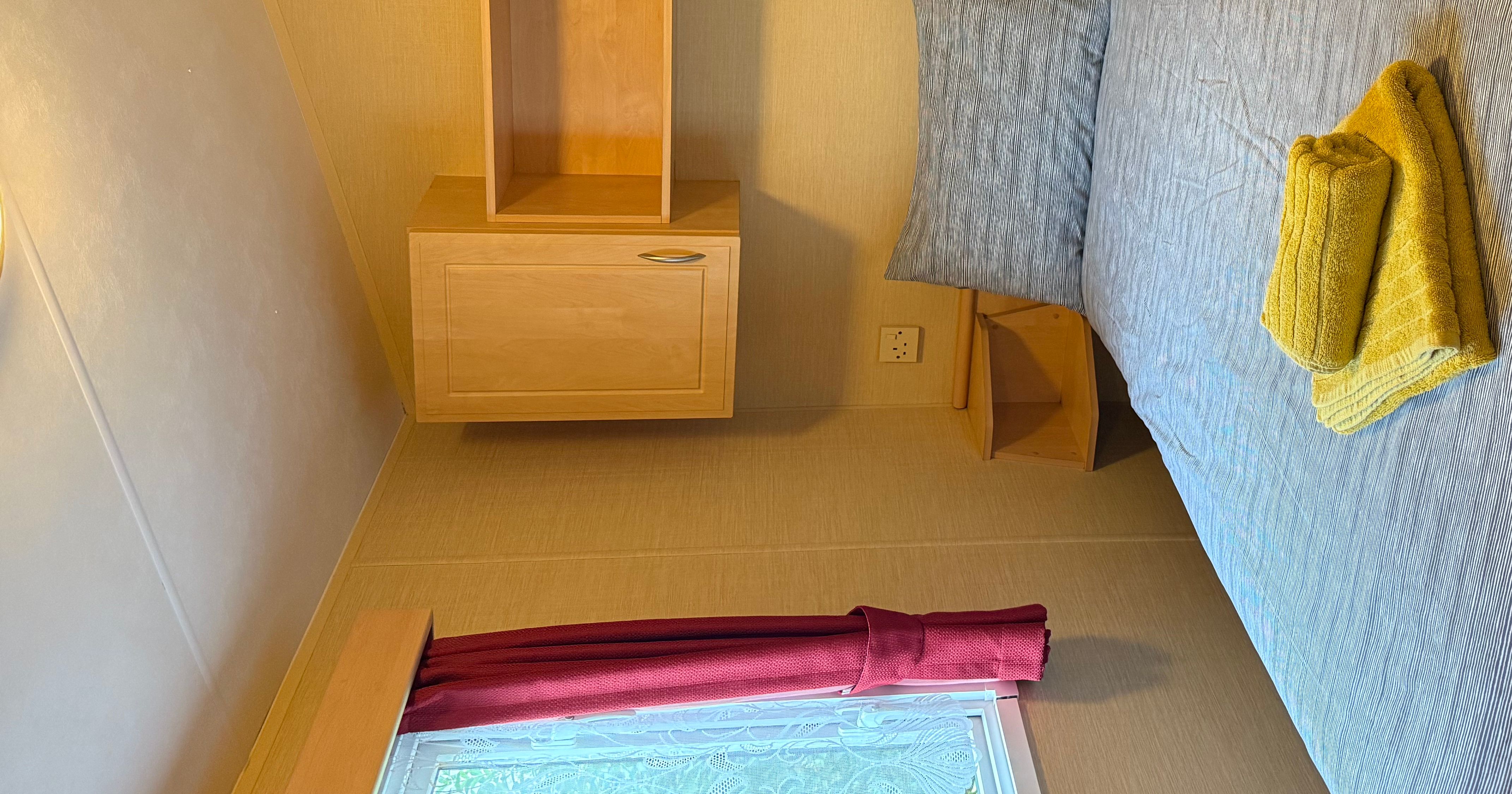 Small bedroom with a double bed, grey bedding, yellow towels, wooden cabinets, and a window with red curtains.