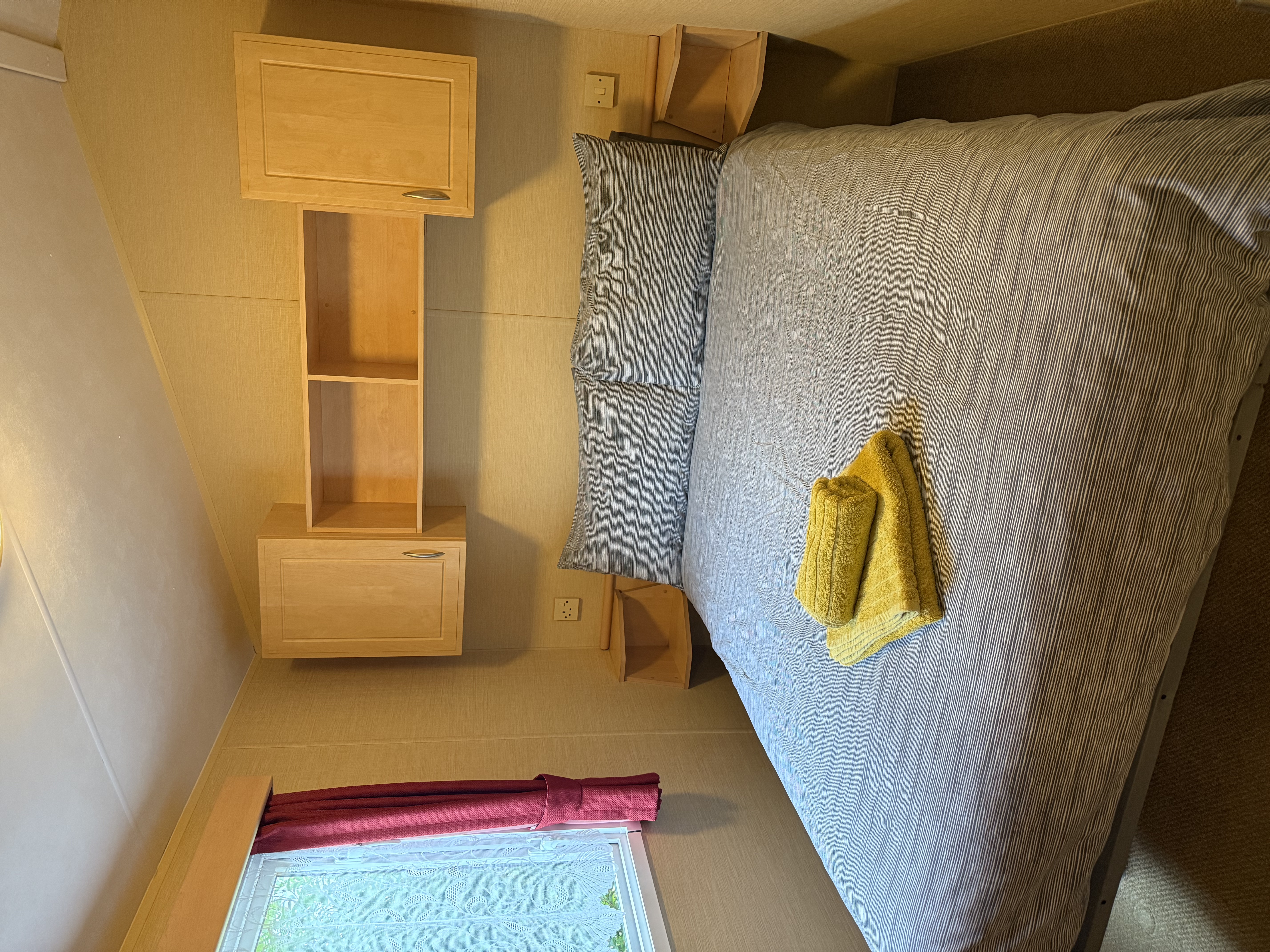 Small bedroom with a double bed, grey bedding, yellow towels, wooden cabinets, and a window with red curtains.