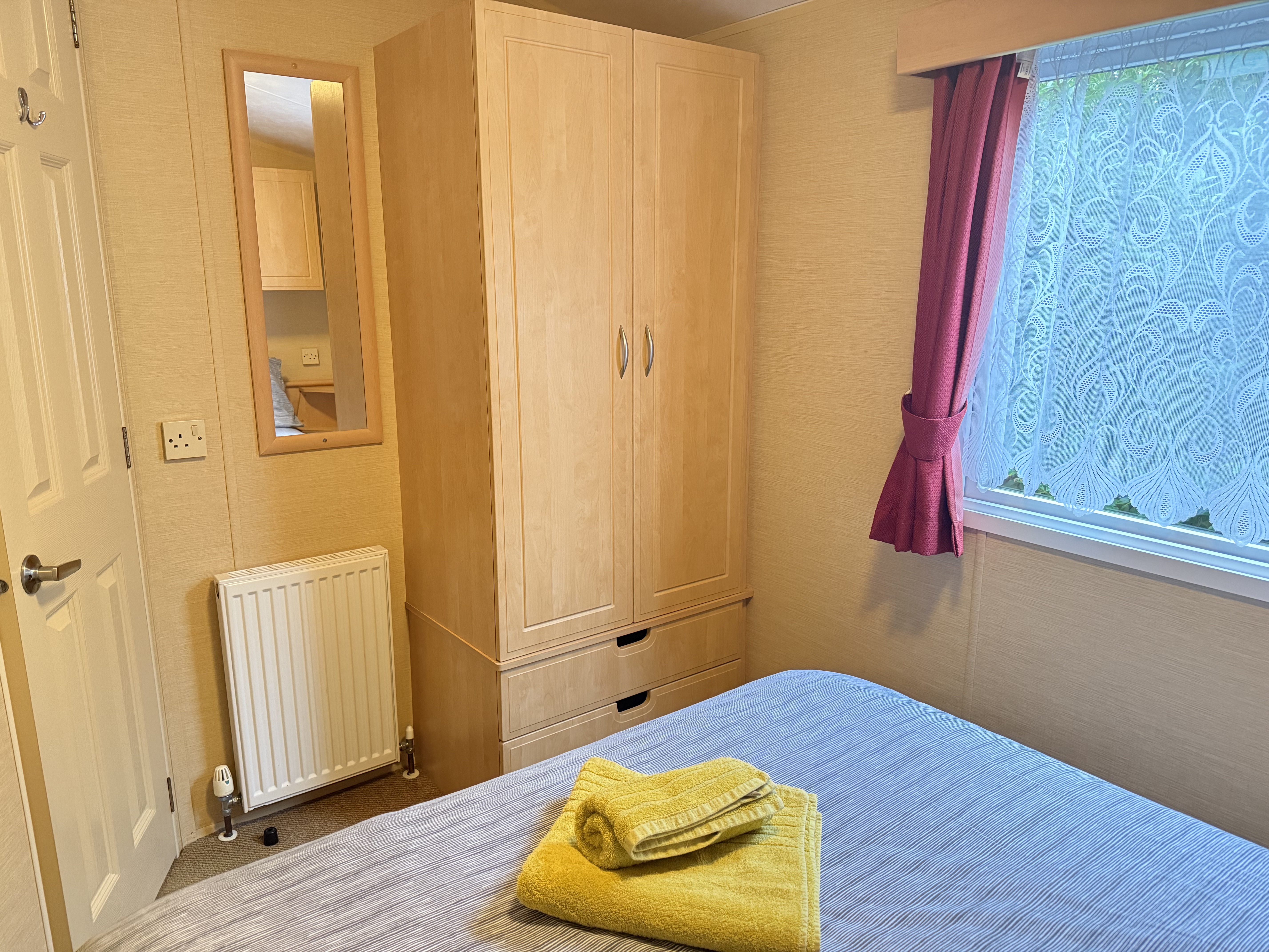 Cozy bedroom with wardrobe, mirror, radiator, and window with red curtains and lacey blinds.
