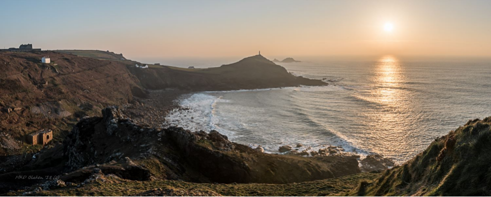 Panoramic view of Cape Cornwall coastline at sunset with the sea, rugged cliffs, and scattered buildings.
