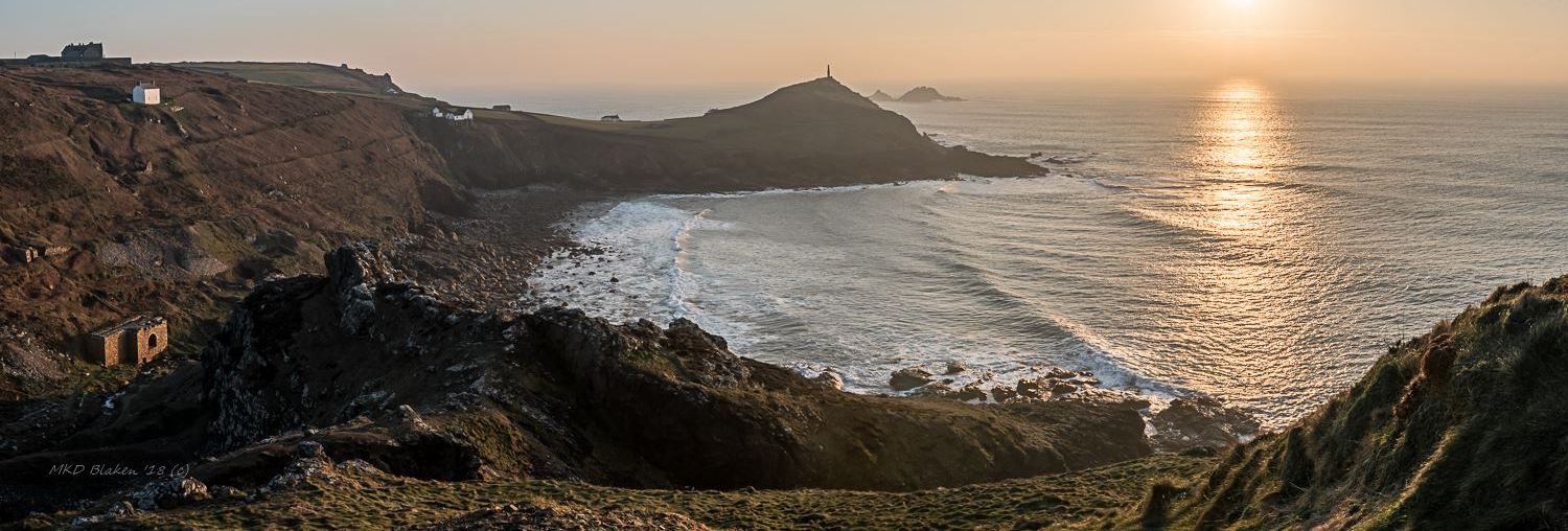 Panoramic view of Cape Cornwall coastline at sunset with the sea, rugged cliffs, and scattered buildings.