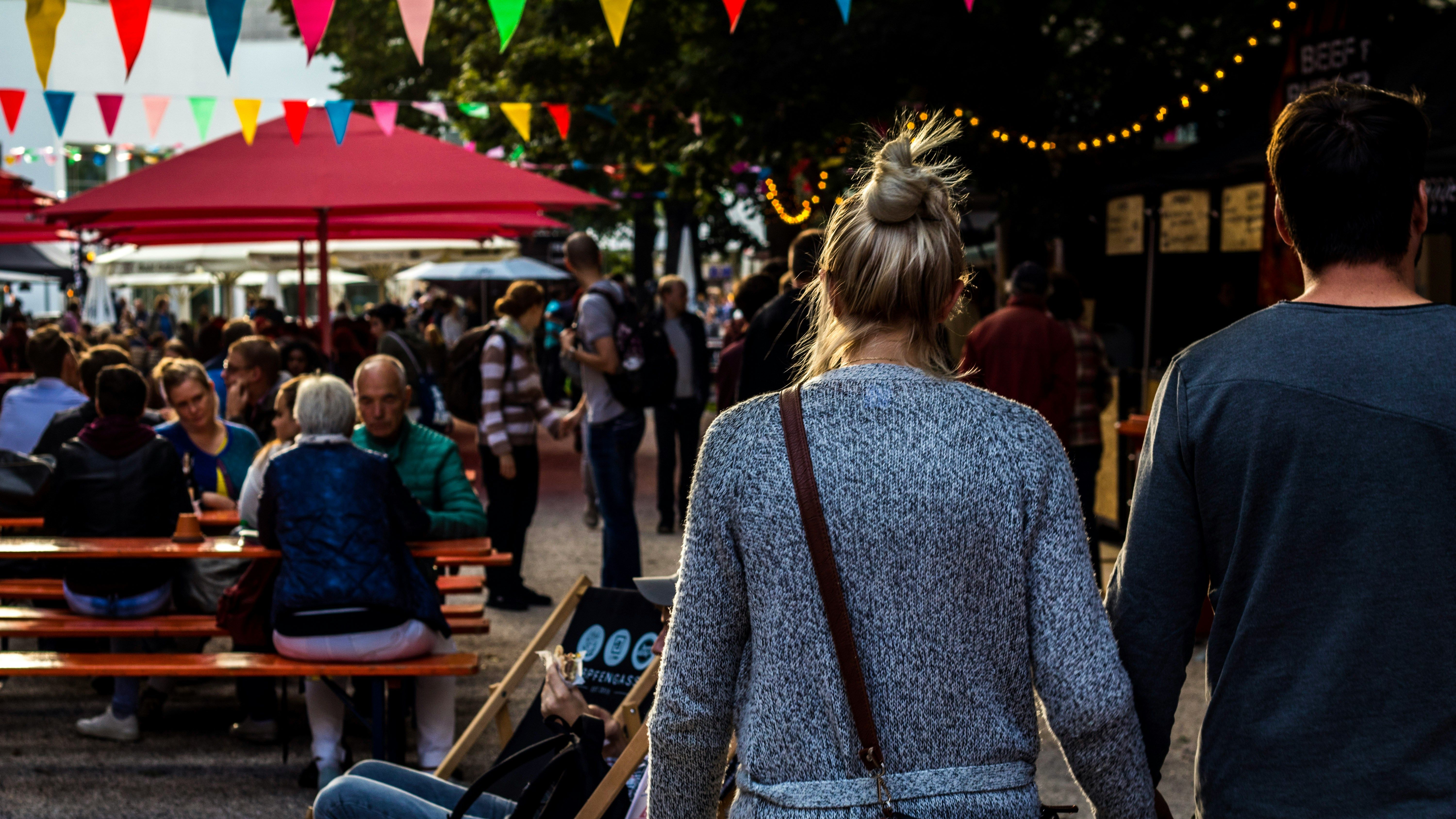Couple holding hands walking through a lively outdoor market or festival with colorful bunting and people sitting at picnic tables.