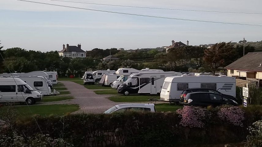 Several caravans and motorhomes parked in an outdoor campsite with a house and trees in the background.