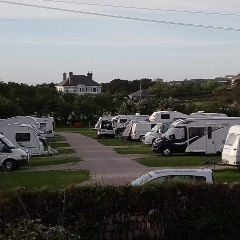 Several caravans and motorhomes parked in an outdoor campsite with a house and trees in the background.