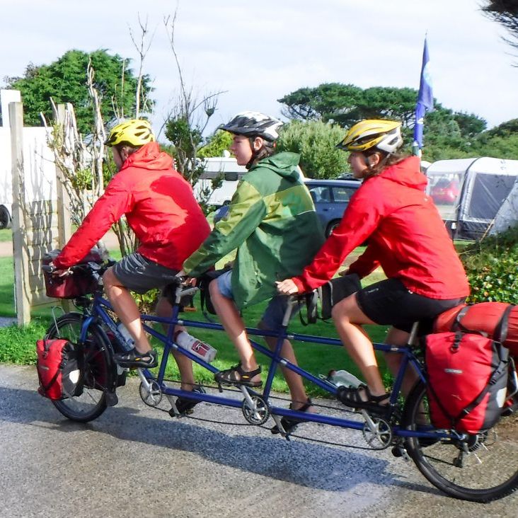 Three people riding a triple tandem bicycle through a campsite with tents and camper vans in the background.