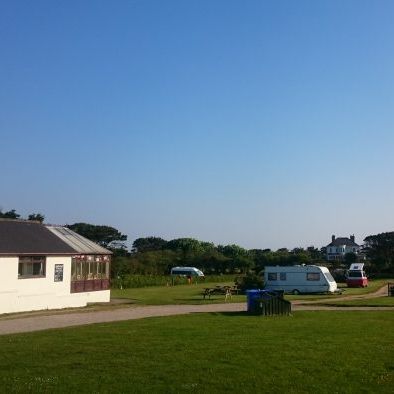 Wide view of a grassy field with a white building, caravans, and distant houses under a clear sky