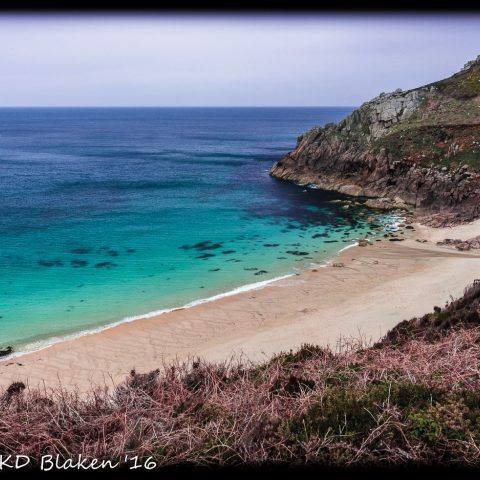 A view of Portheras Cove from a path above, showing turquoise waters, sandy beach, and rocky hillside.