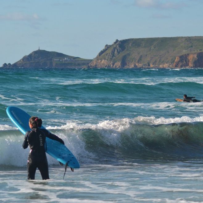 A person in a wetsuit holding a blue surfboard stands in the waves, with another surfer paddling in the ocean and cliffs in the background.