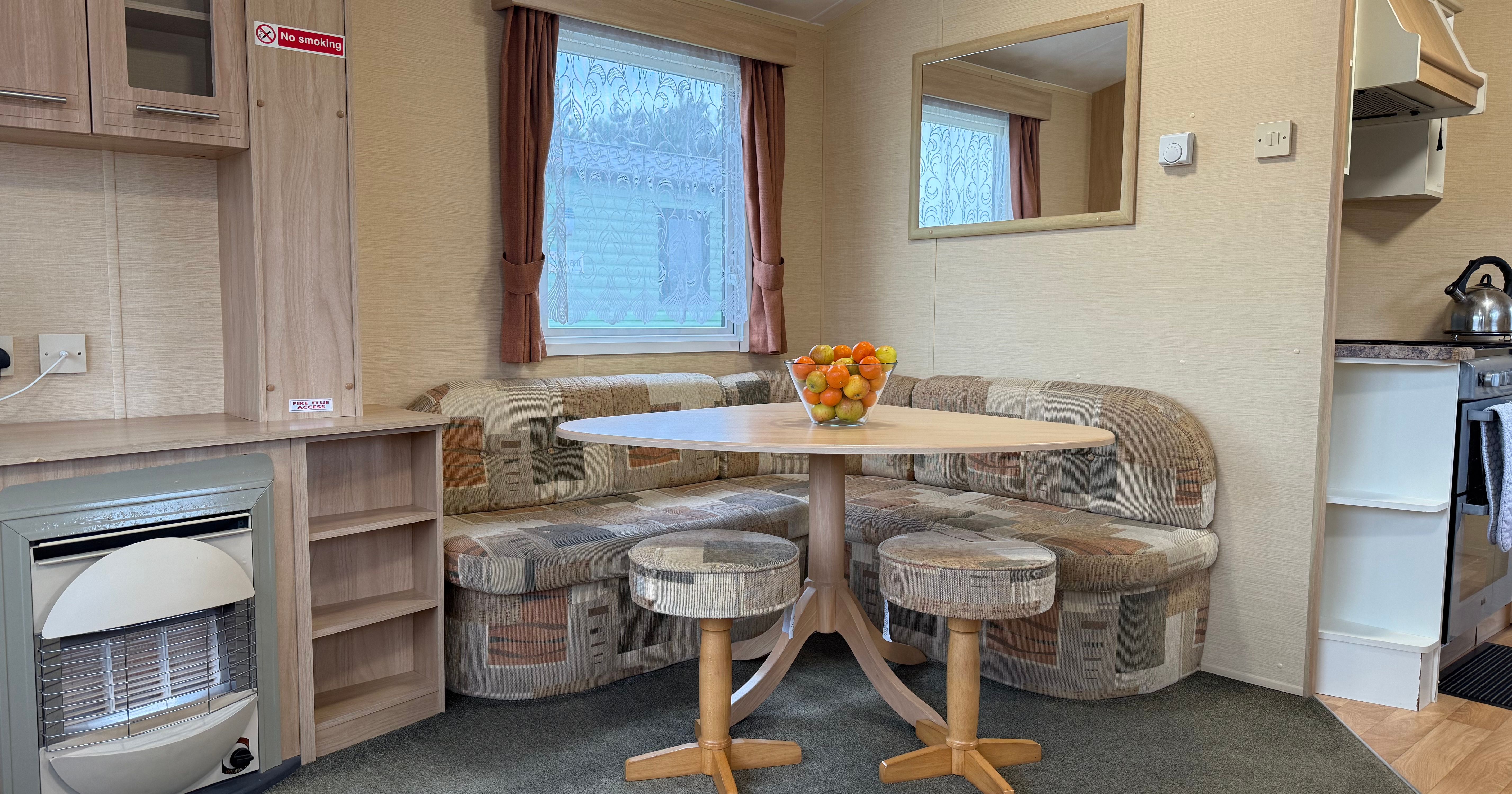 Interior of a cozy caravan or mobile home dining area with a round table, cushioned bench seating, and a bowl of fruit.