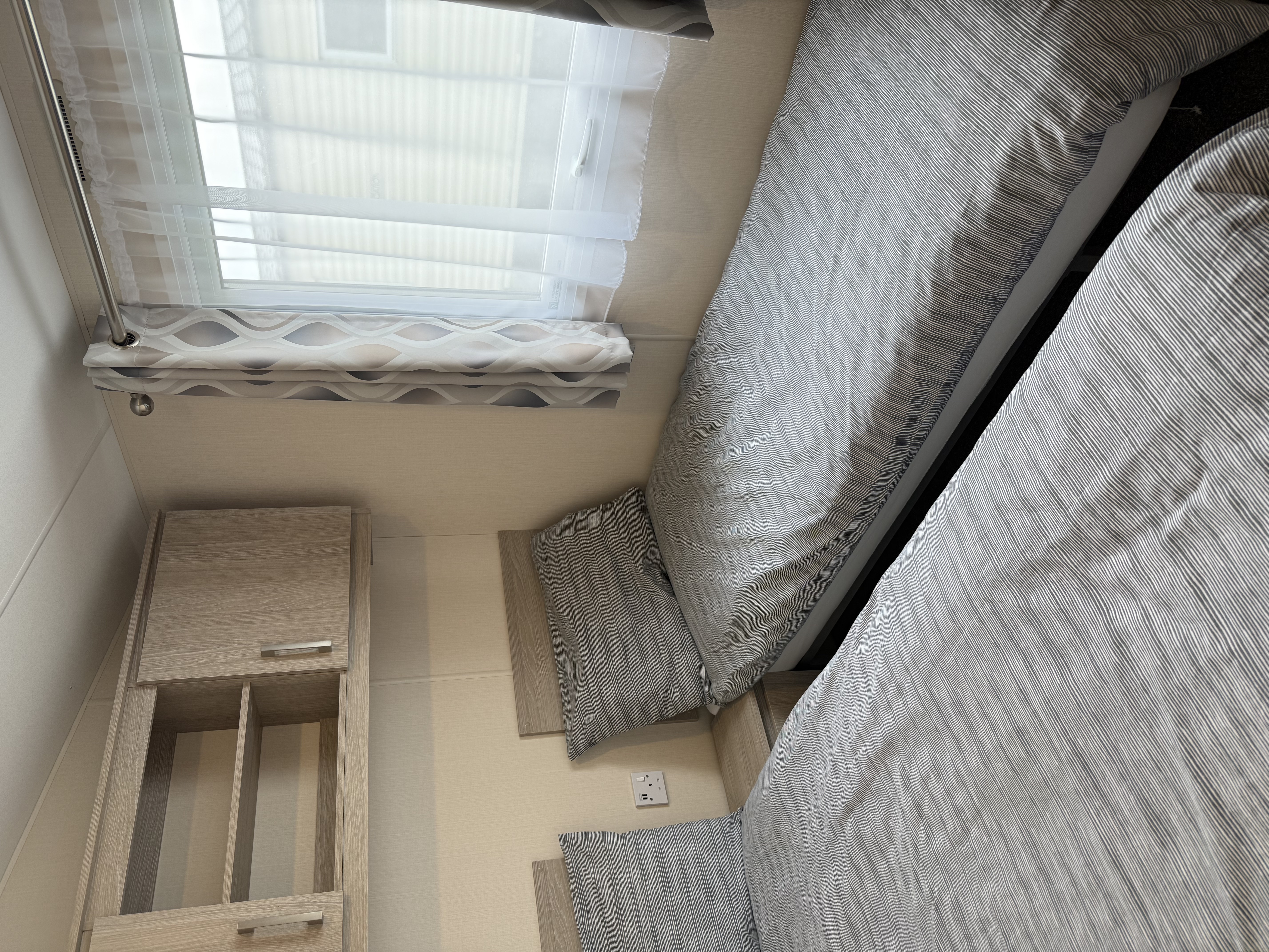 Small bedroom with two single beds, grey striped bedding, a window with patterned curtains, and wooden shelving above.