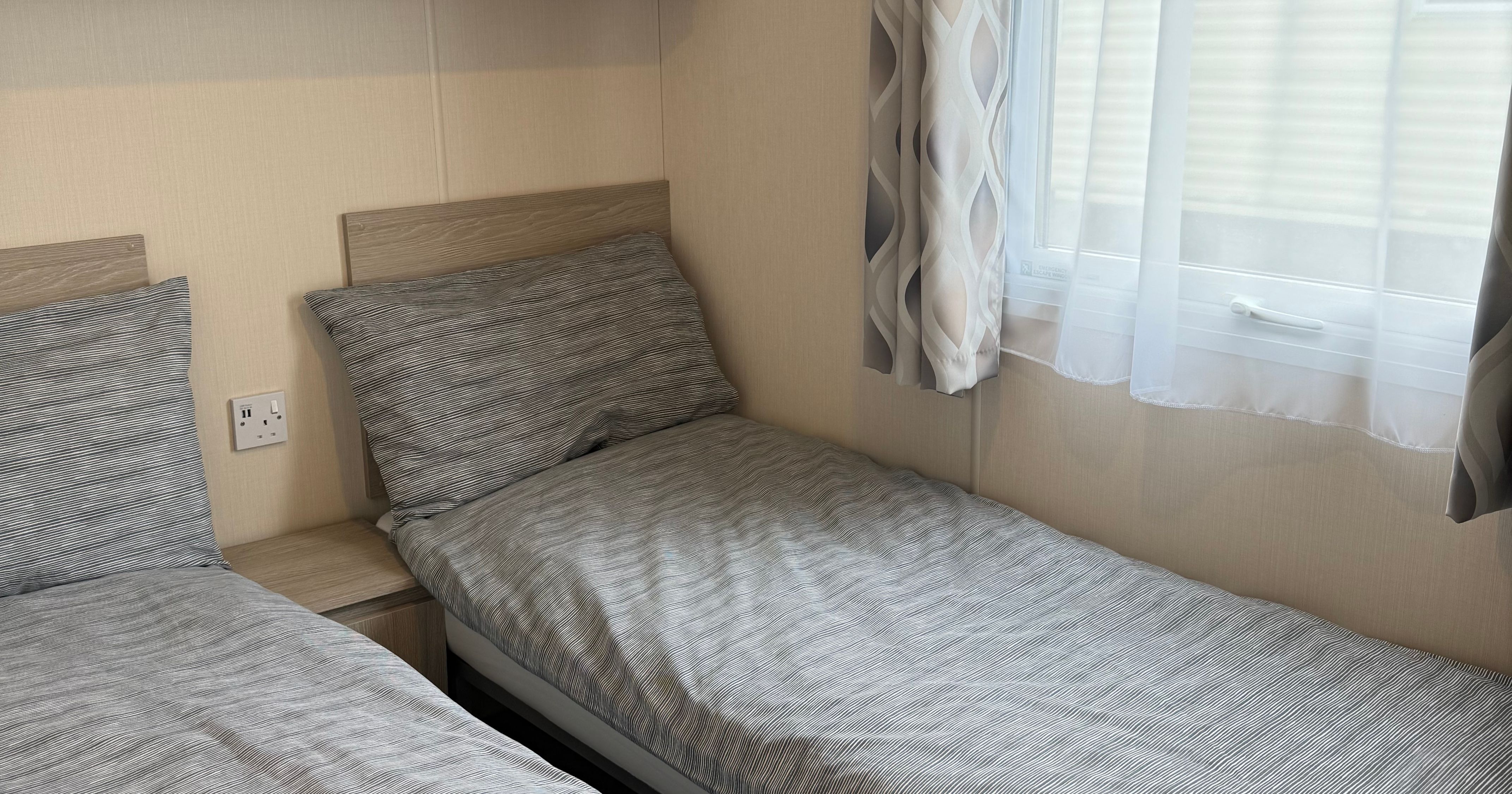 Small bedroom with two single beds, grey striped bedding, a window with patterned curtains, and wooden shelving above.