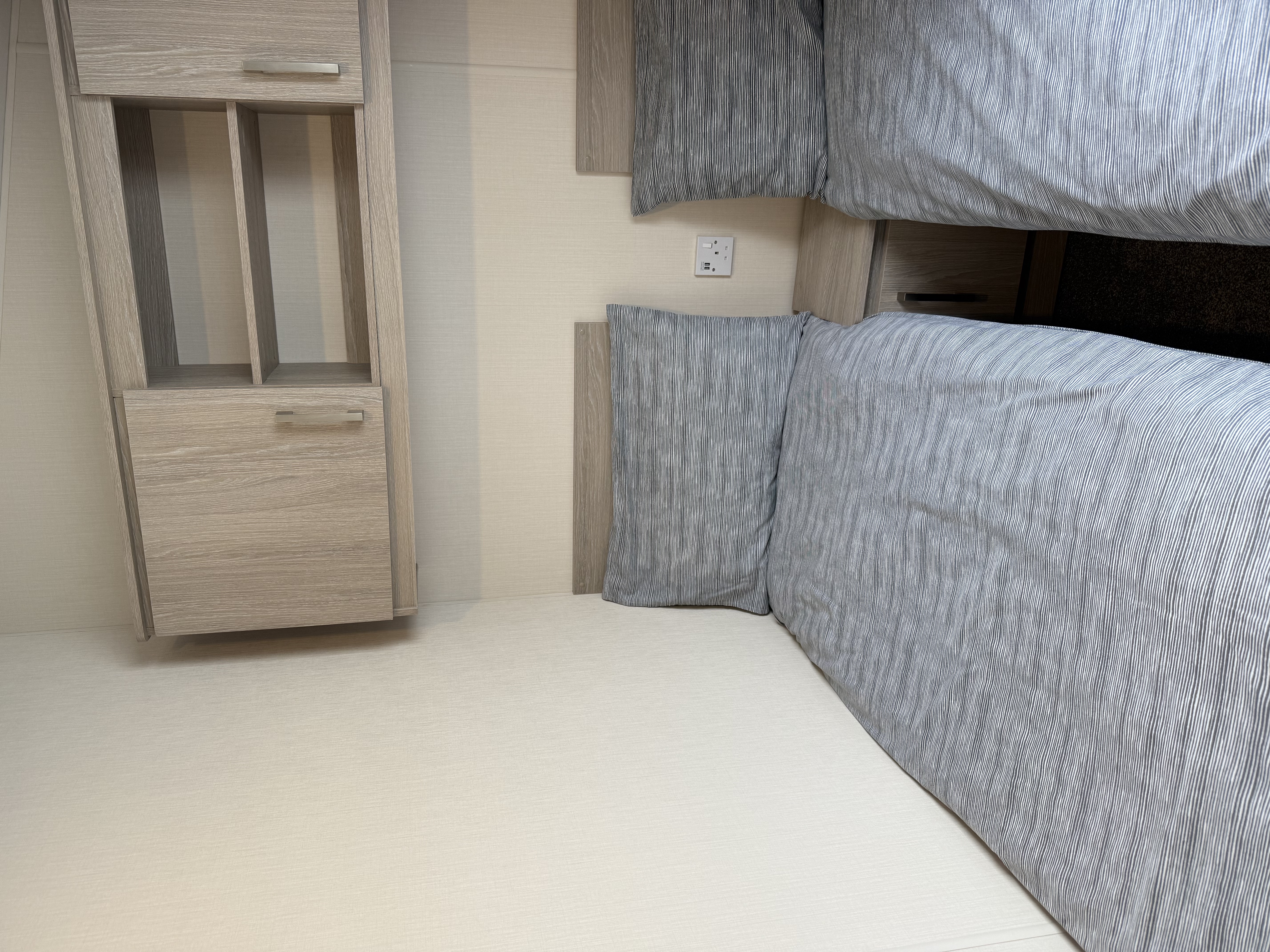 Two single beds with grey bedding next to a small nightstand and wooden shelves above.