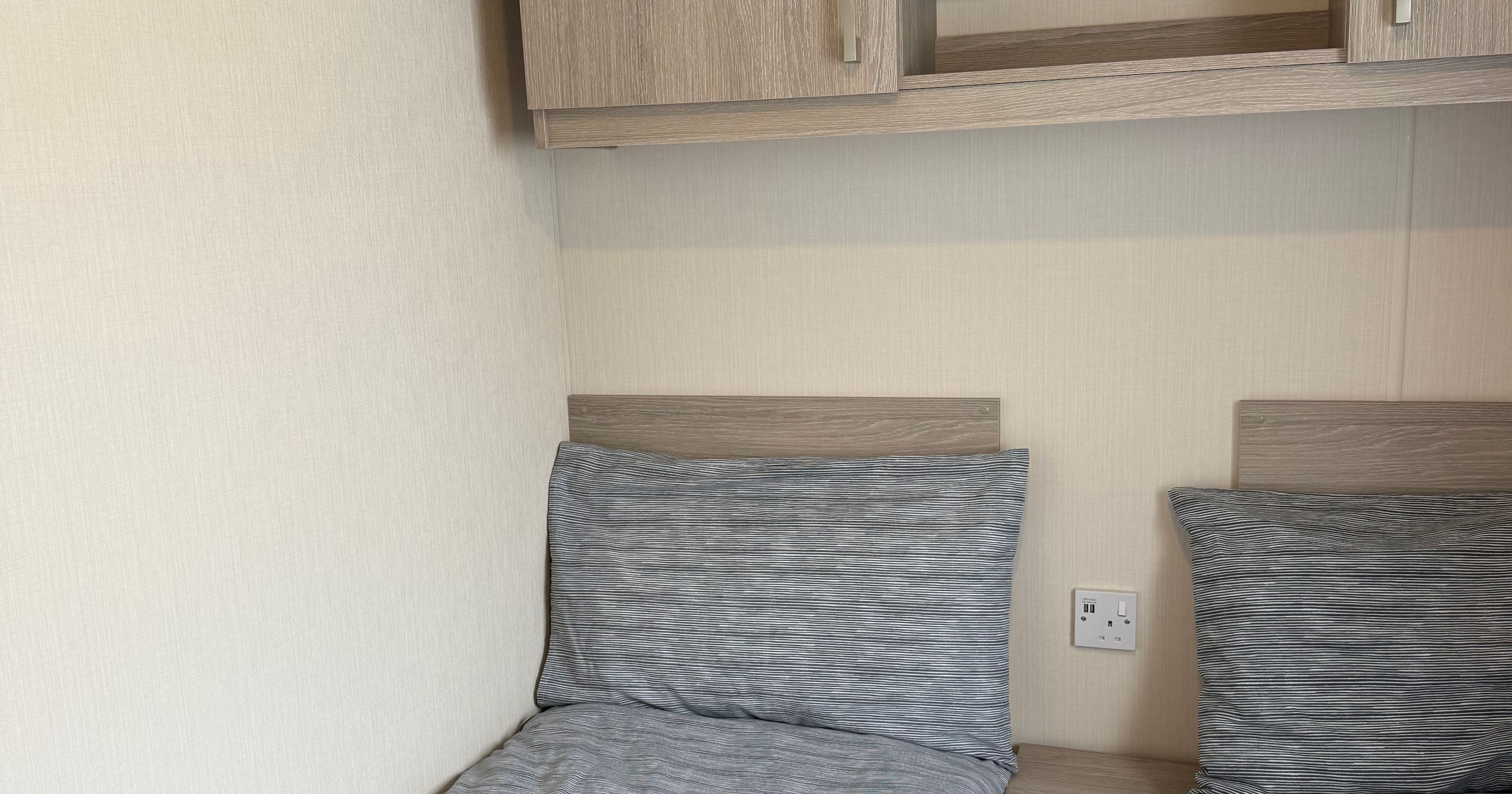 Two single beds with grey bedding next to a small nightstand and wooden shelves above.