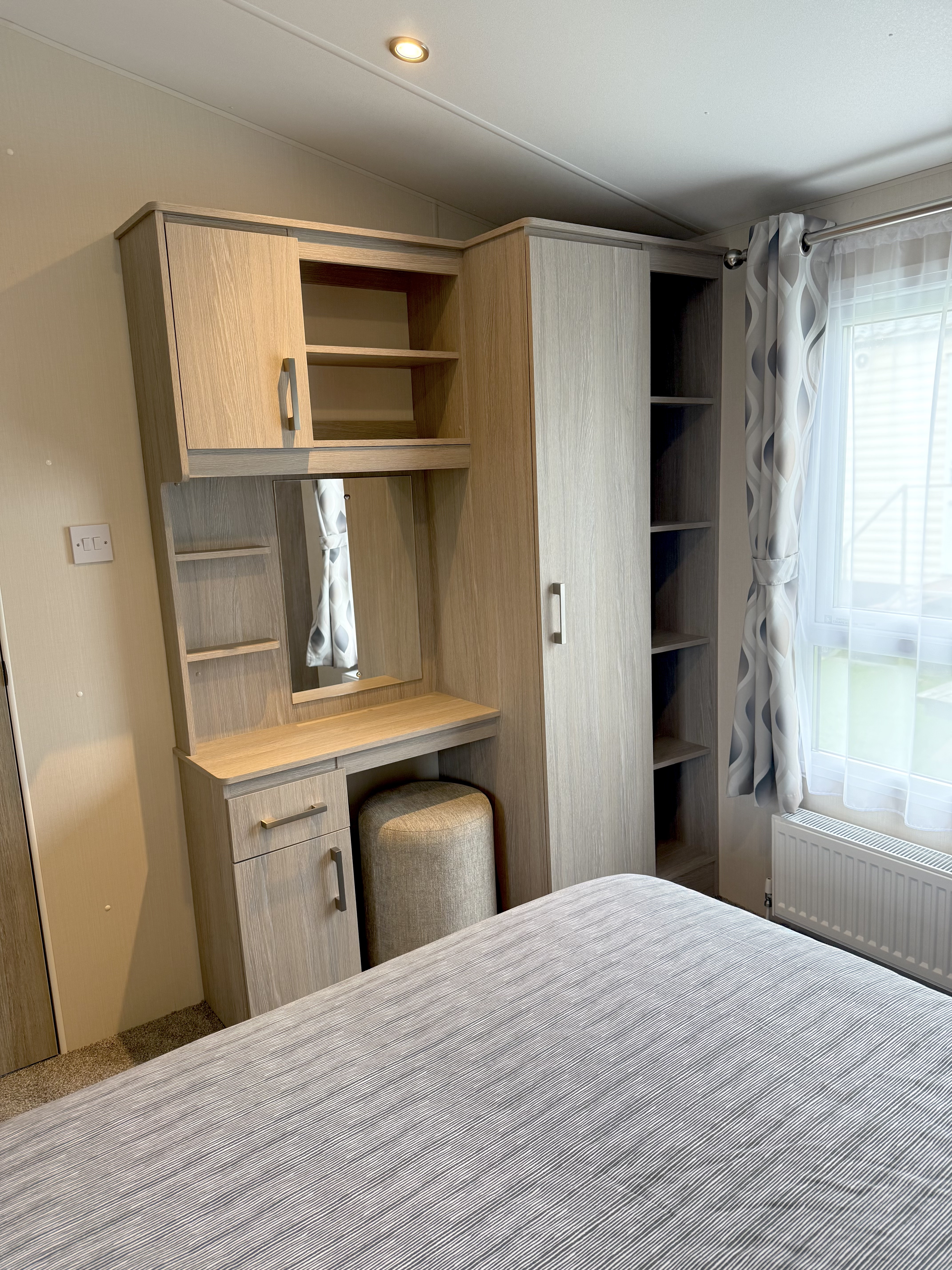 A modern wooden vanity and wardrobe set in a bedroom with a grey bedspread and window with patterned curtains.