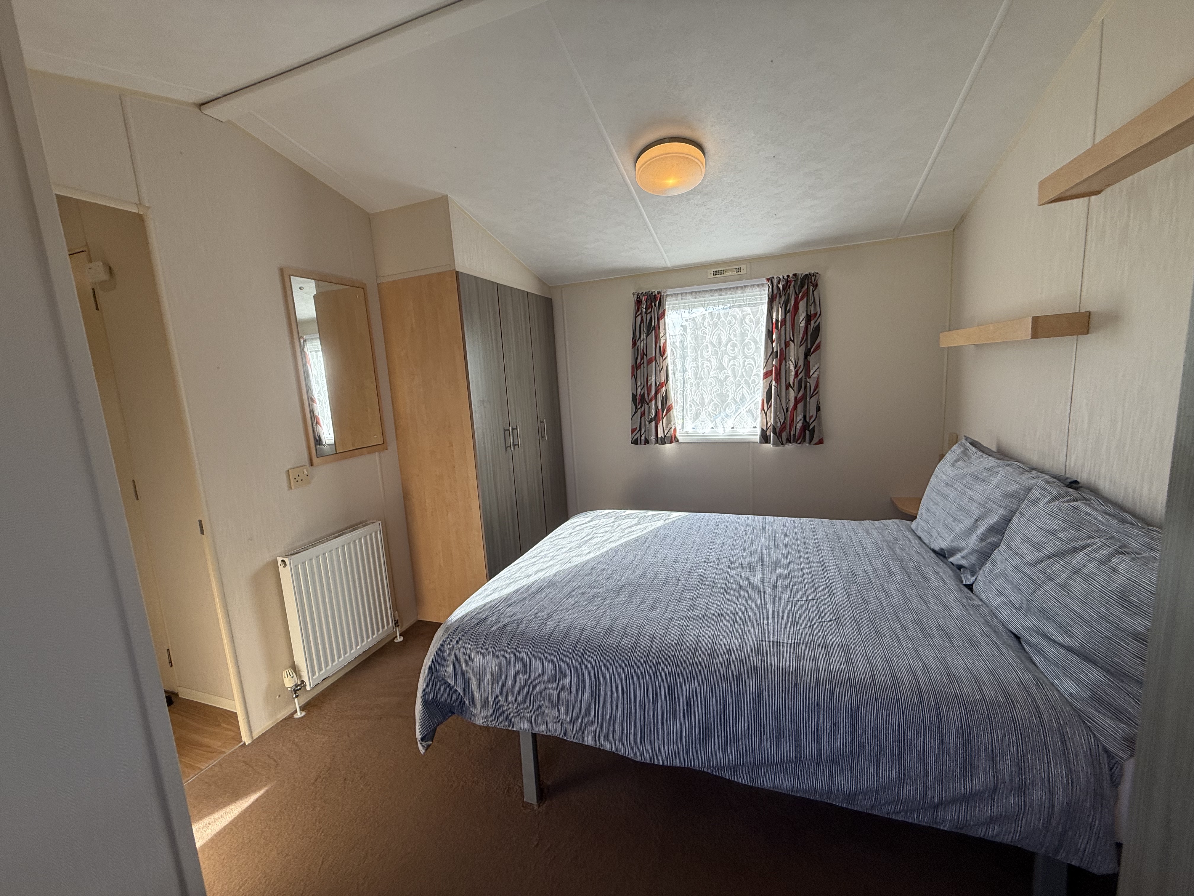 A tidy bedroom with a double bed, wardrobe, mirror, and a window with patterned curtains.