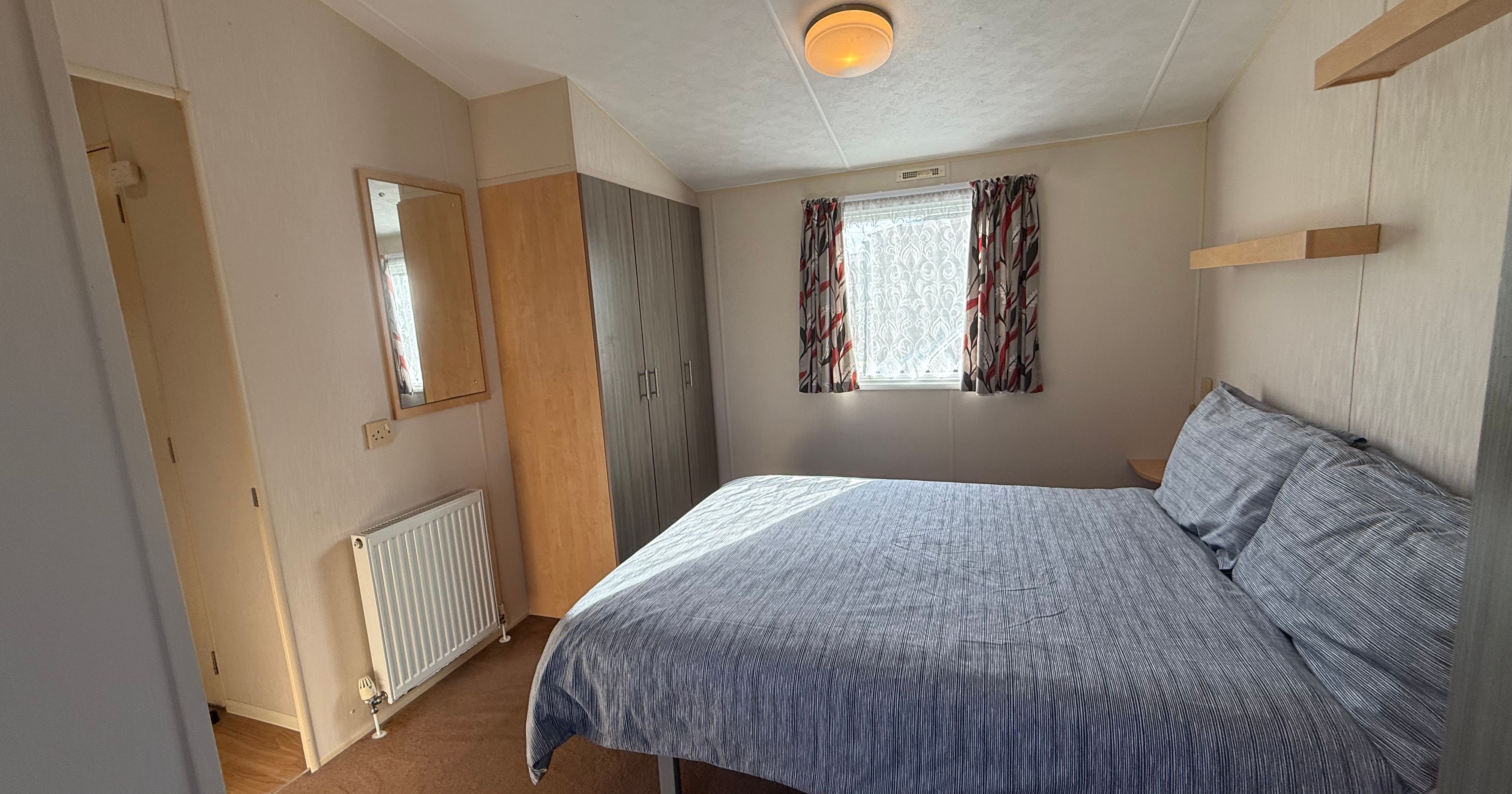 A tidy bedroom with a double bed, wardrobe, mirror, and a window with patterned curtains.