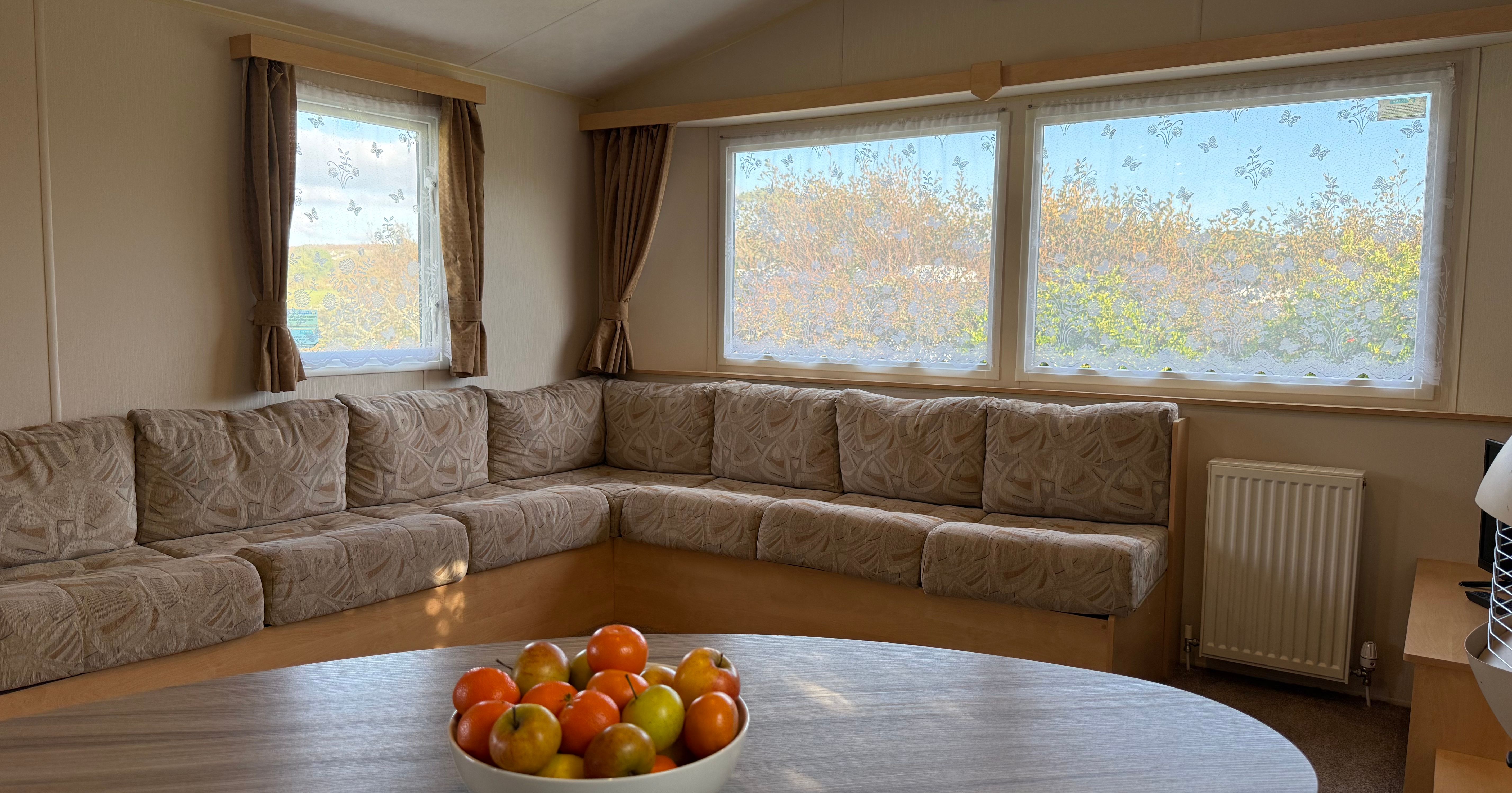 Interior of a cozy living room with beige sofa, large windows with lace curtains, and a bowl of fruit on a table.