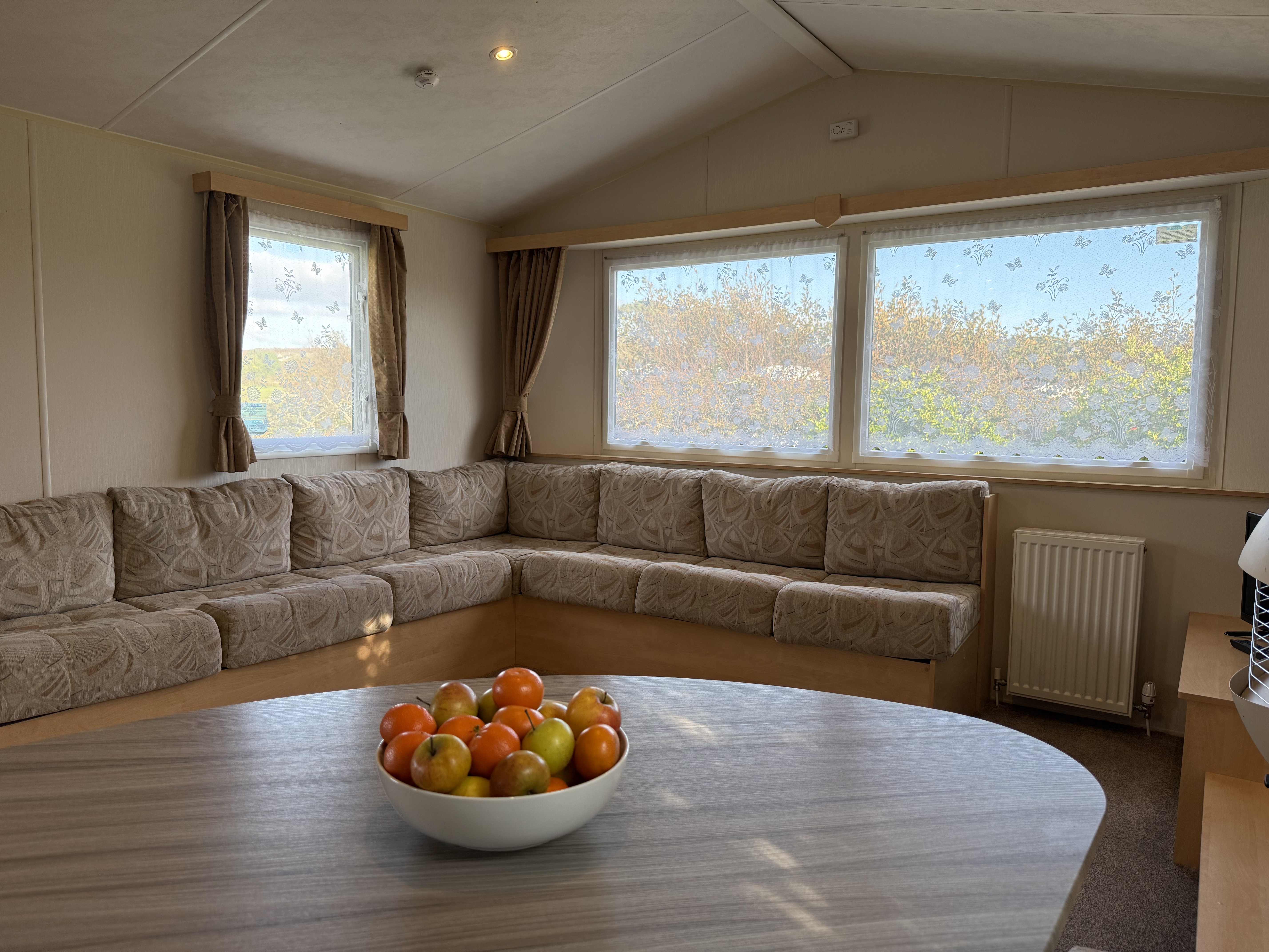 Interior of a cozy living room with beige sofa, large windows with lace curtains, and a bowl of fruit on a table.