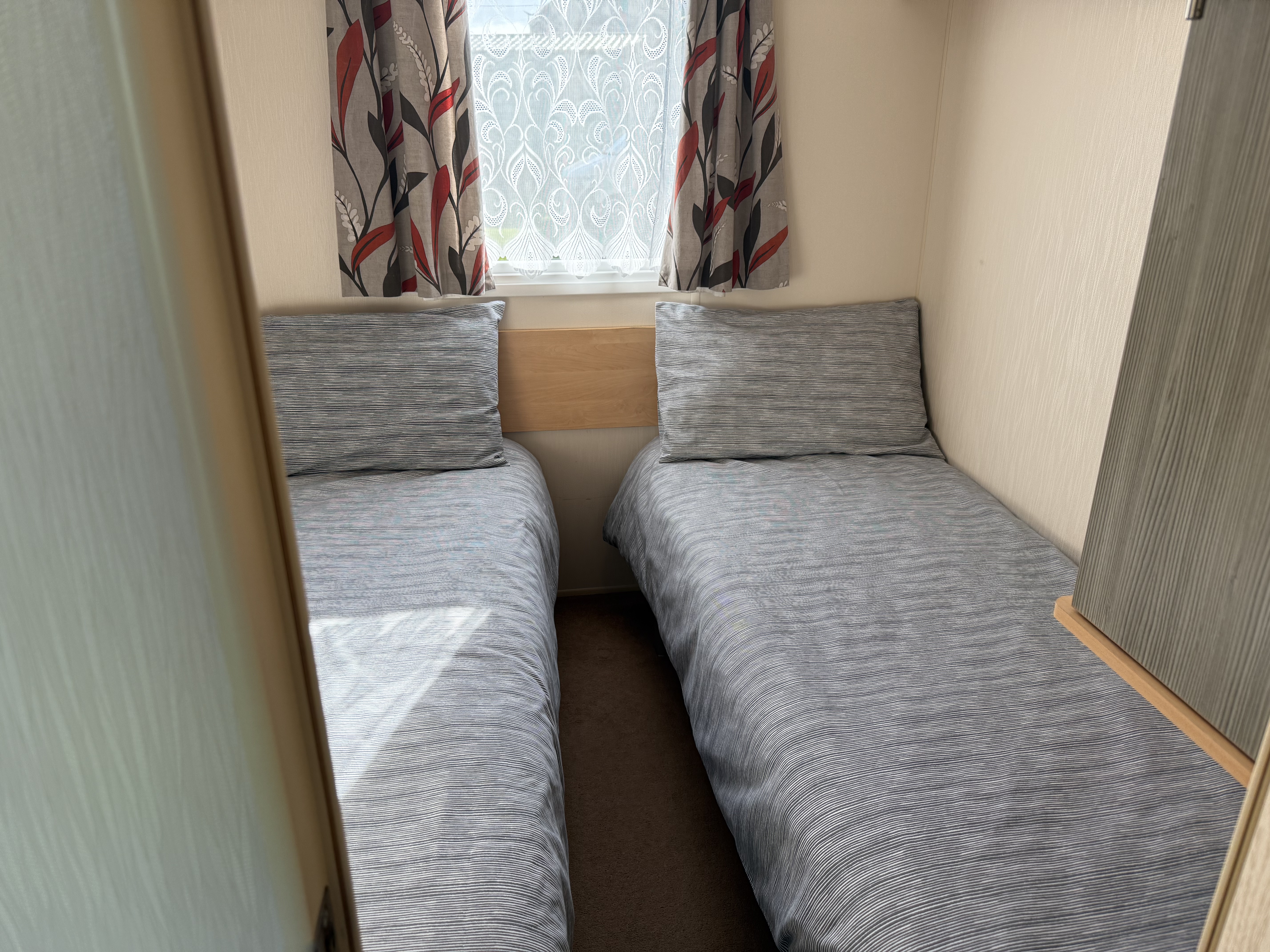 A small bedroom with two single beds, each with grey bedding and a large pillow. A window with patterned curtains is at the head of the beds.