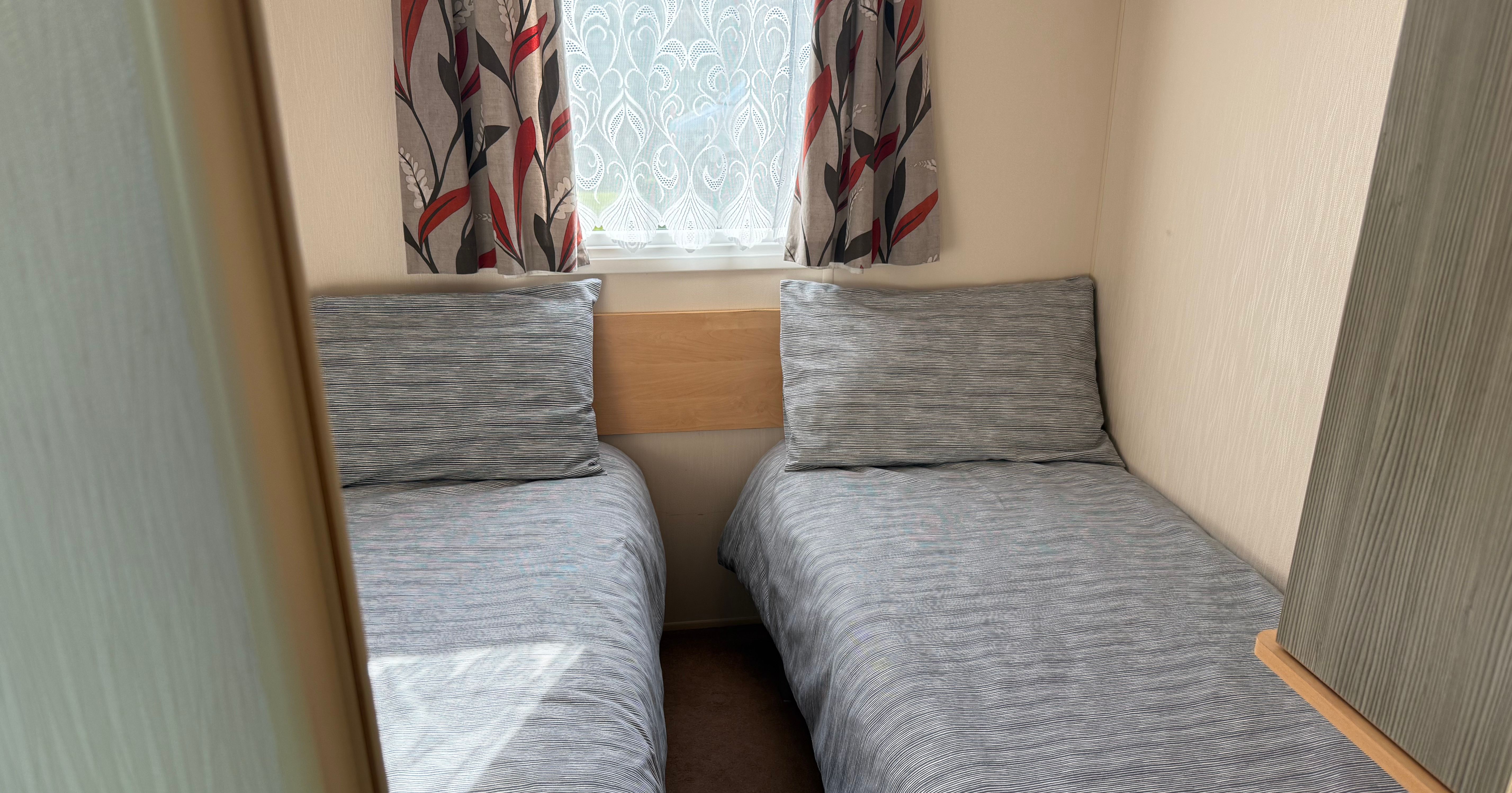 A small bedroom with two single beds, each with grey bedding and a large pillow. A window with patterned curtains is at the head of the beds.