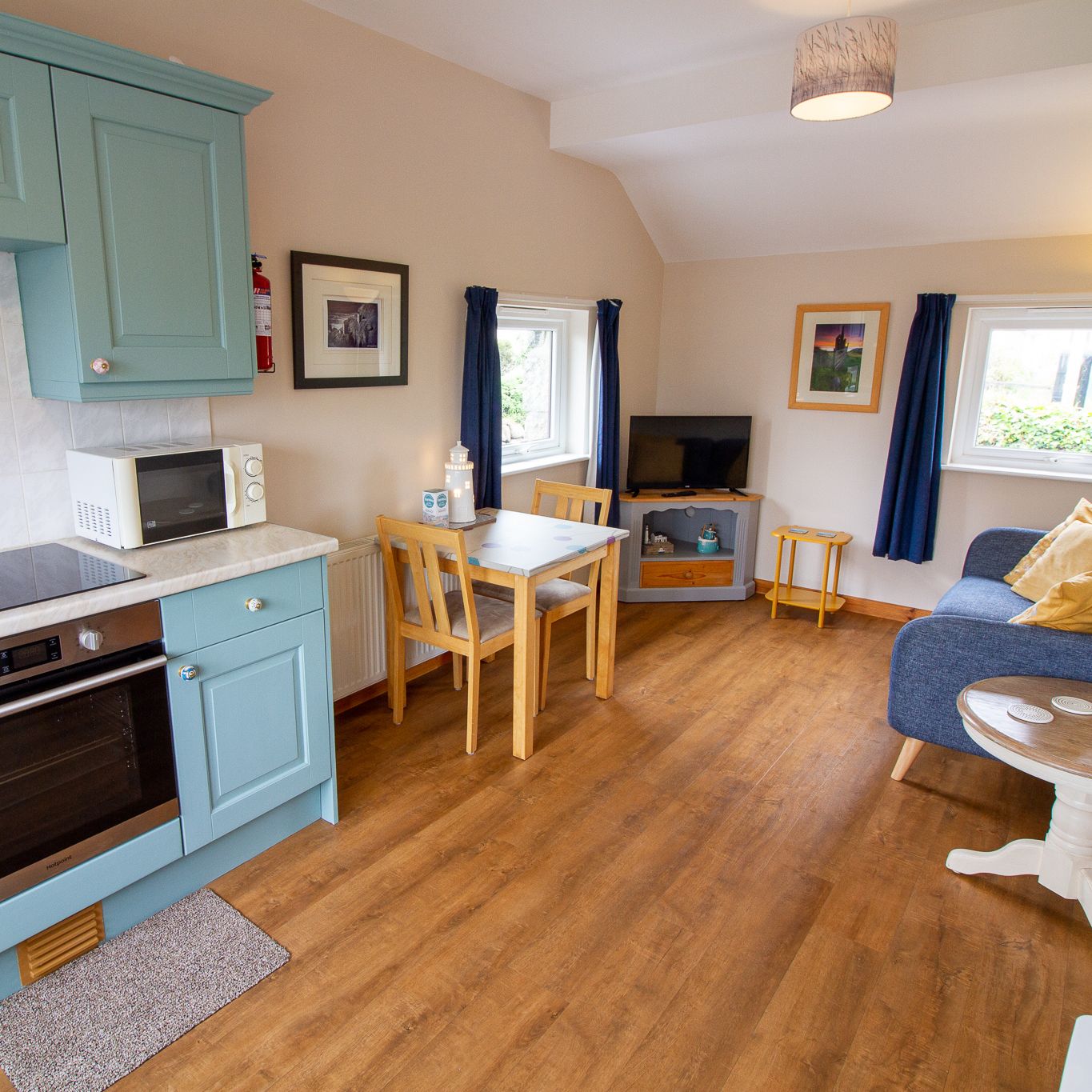 Cozy open-plan kitchen and living area with blue cabinets, small dining table, sofa, and TV.