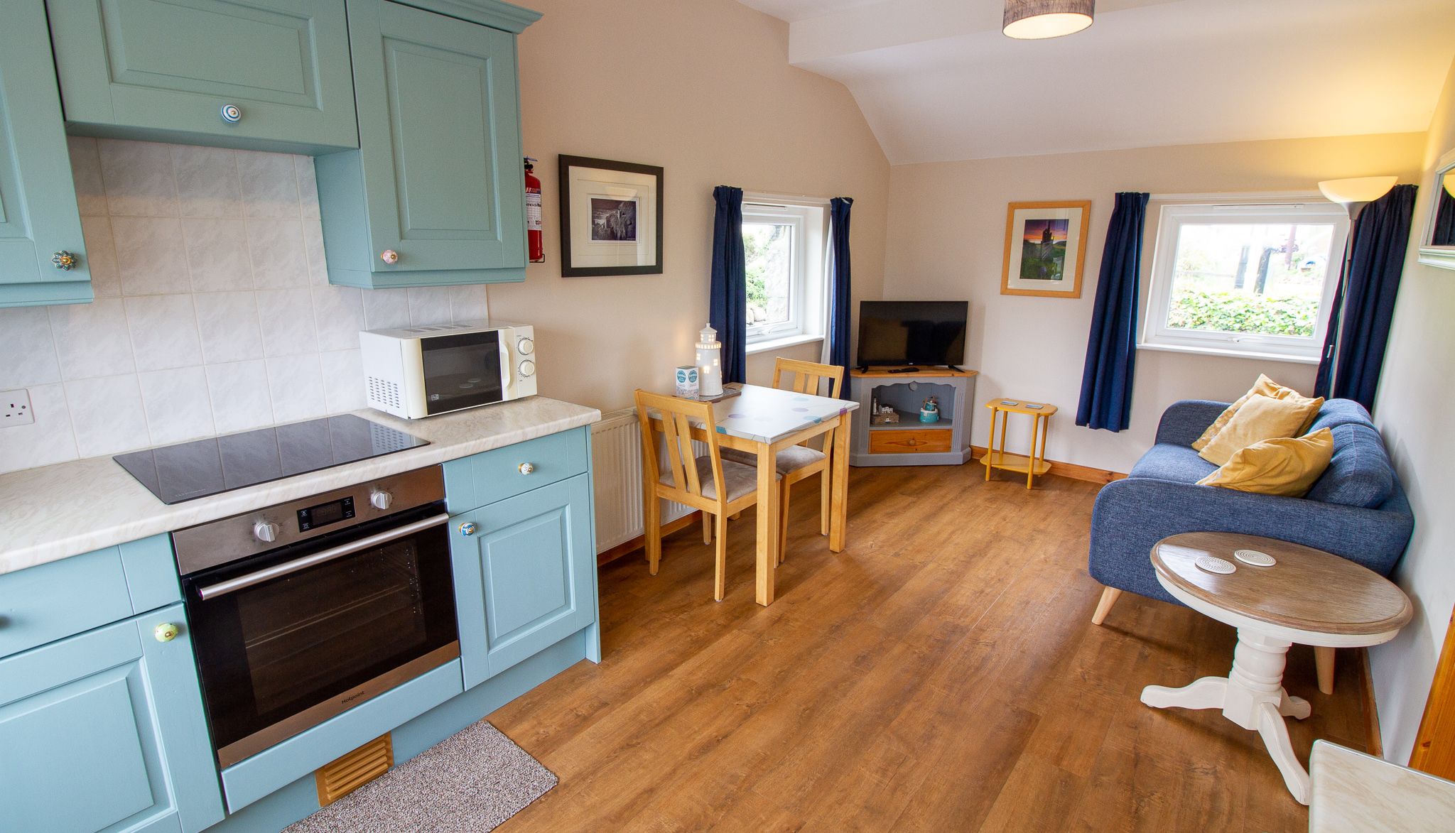 Cozy open-plan kitchen and living area with blue cabinets, small dining table, sofa, and TV.